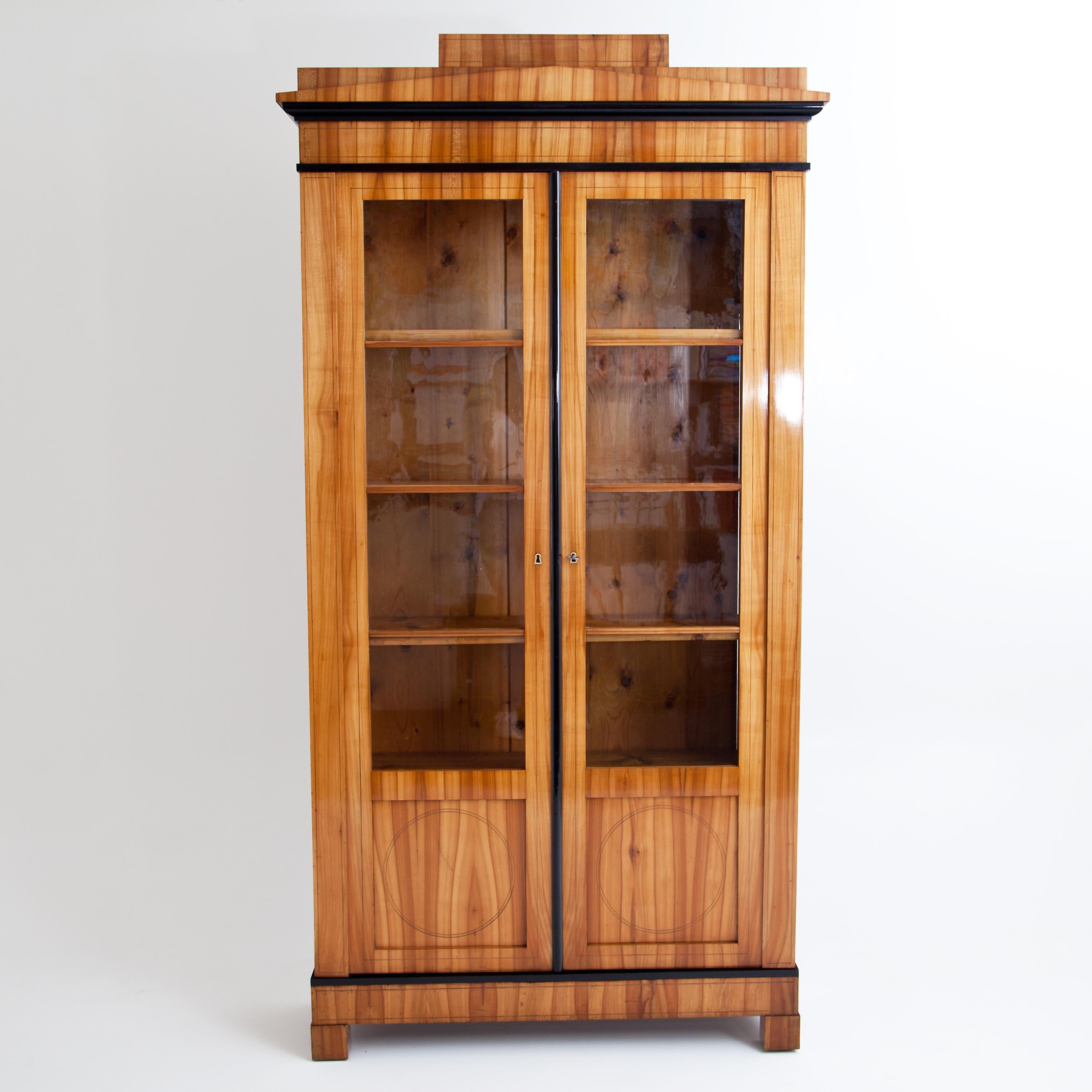 Two-door bookcase on low square feet with doors in 4/5 glazing and stepped cornice. The plinth and cornice are optically structured by ebonized profiles and harmoniously united by the vertical ebonized strip. The corpus is veneered in cherrywood and