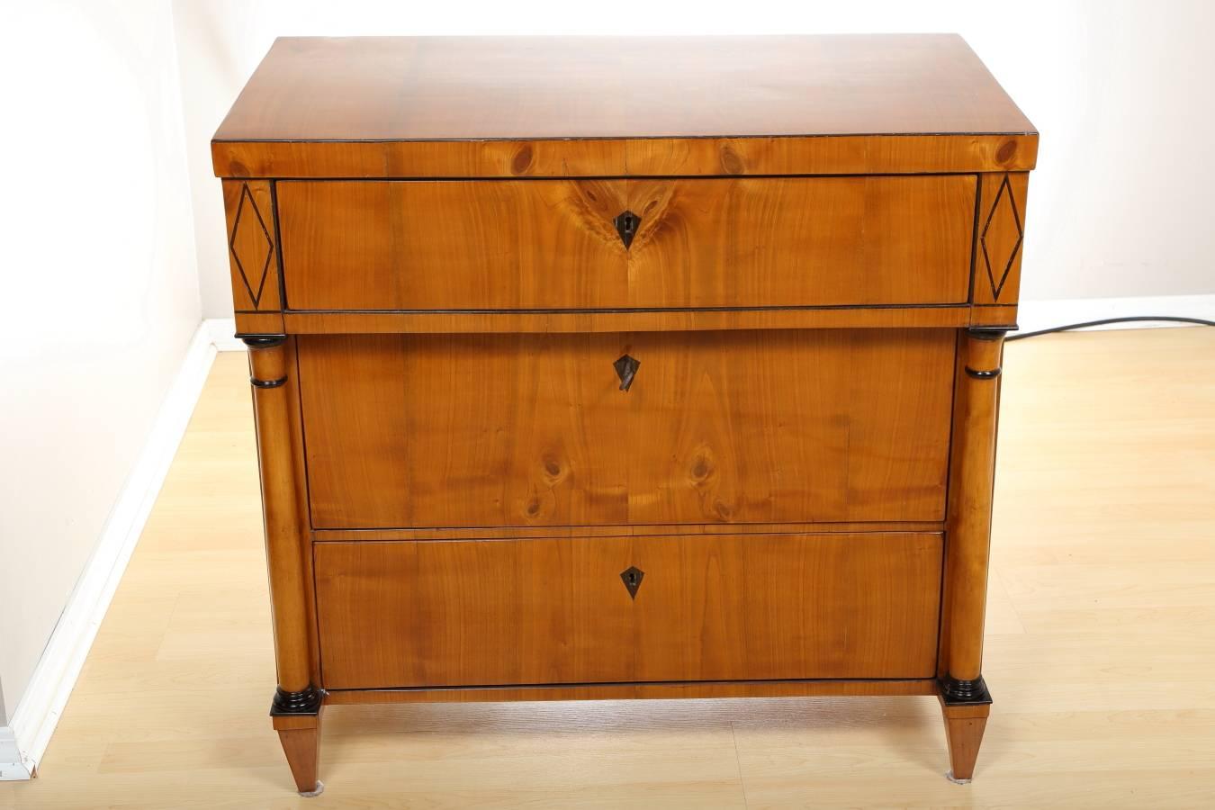 Biedermeier cherry commode, circa 1840. This elegant cherry chest of drawers features three large drawers flanked by round columns resting on tapered feet. Ebonized wood diamond shape inlays and column details add to the beauty of the piece. The