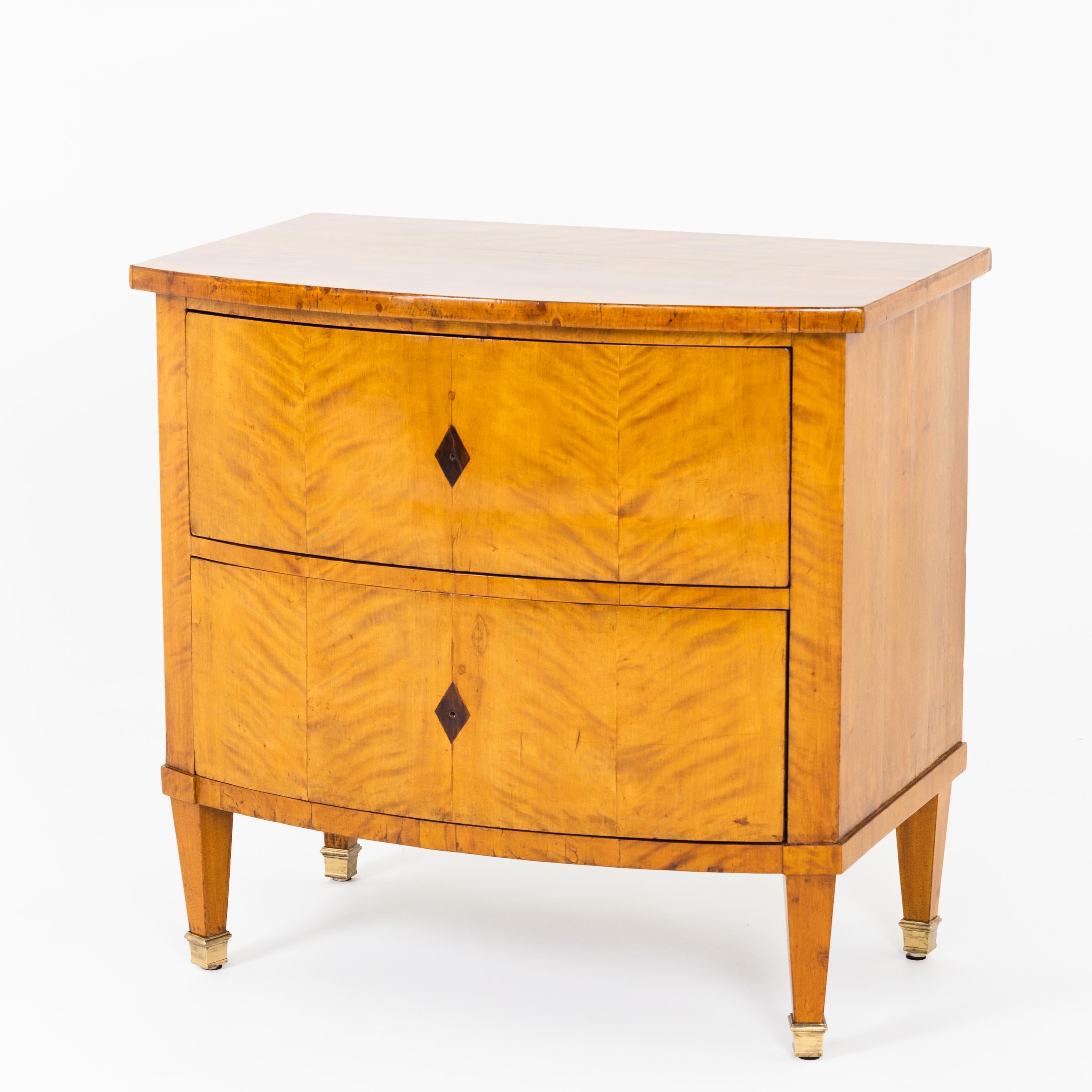 A Biedermeier chest of drawers standing on square tapering feet with brass sabots, with a bow front and two drawers with diamond-shaped inlays. The corpus is veneered in birch.
  
