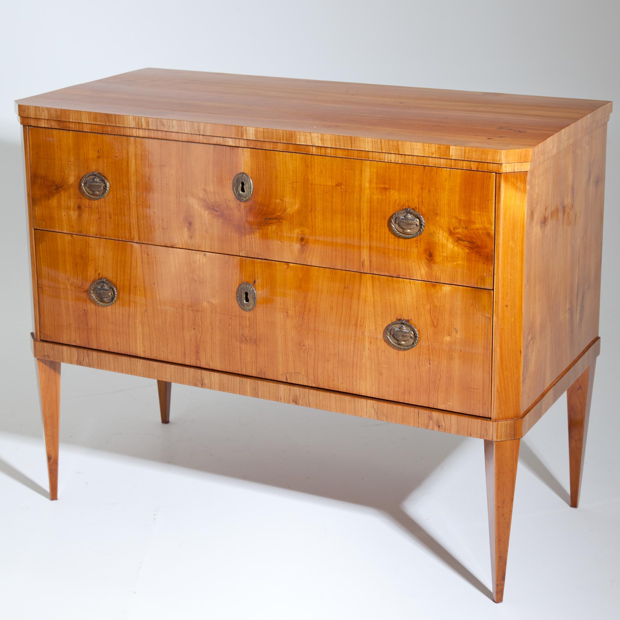 Two-drawered chest of drawers with bevelled corners, veneered in cherry, standing on tall tapered feet. Brass fittings. Expertly restored condition.