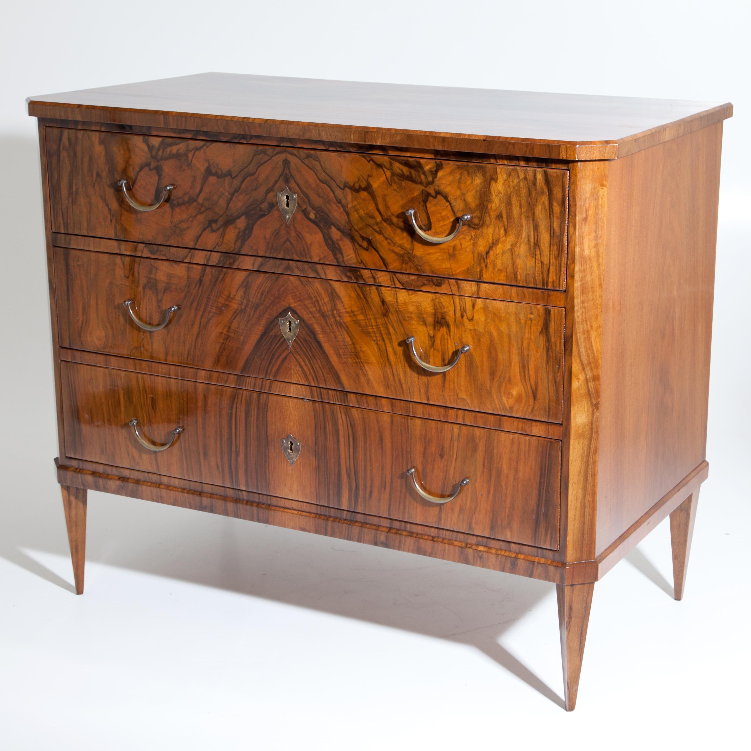 Chest of drawers standing on square pointed feet with three drawers and bevelled corners. The beautiful walnut veneer is mirrored on the front. Expertly restored condition. Fittings are recent.