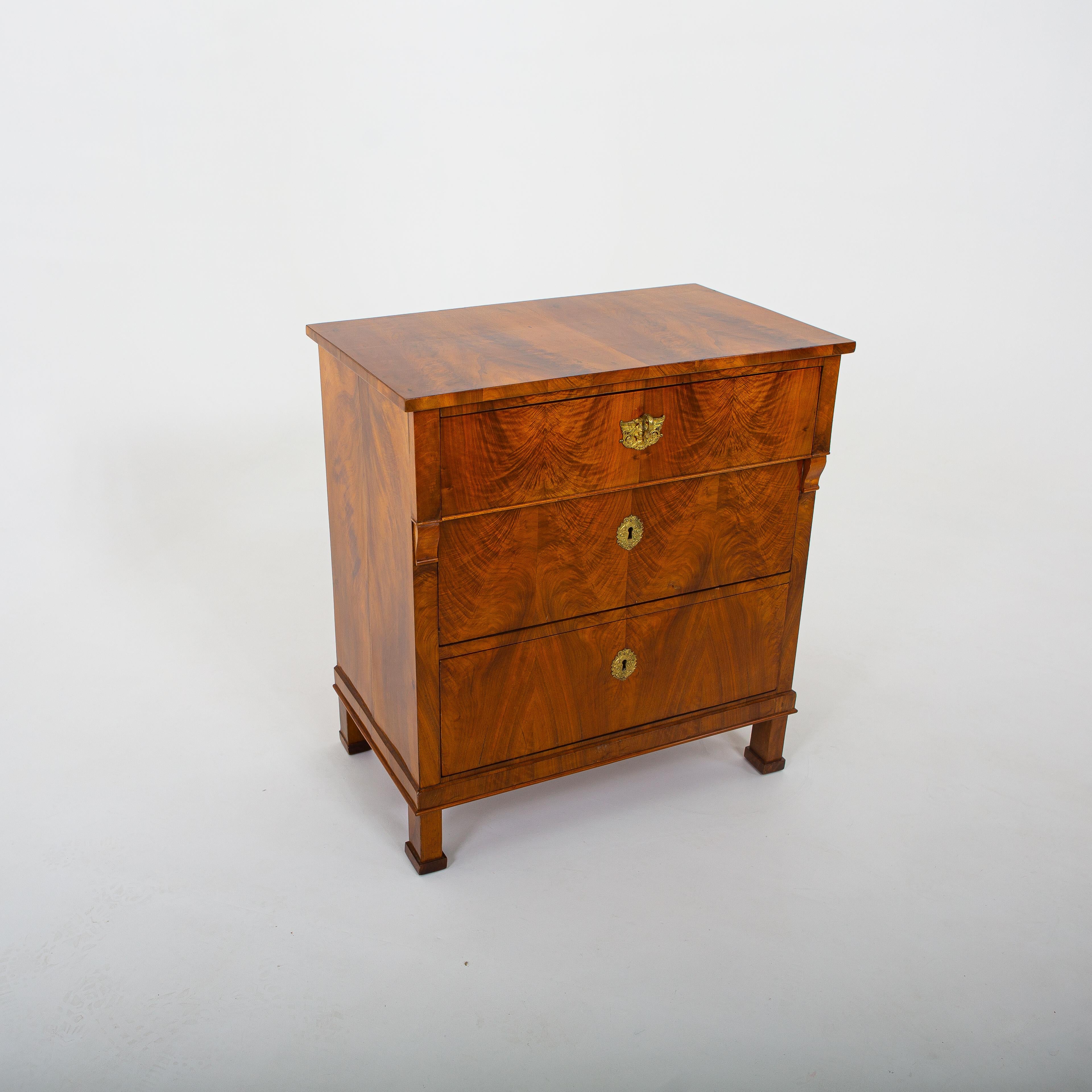 Biedermeier chest of drawers on square feet, veneered in walnut, with three drawers and gilded key escutcheons and fittings. The top drawer protrudes slightly and rests on small volutes.