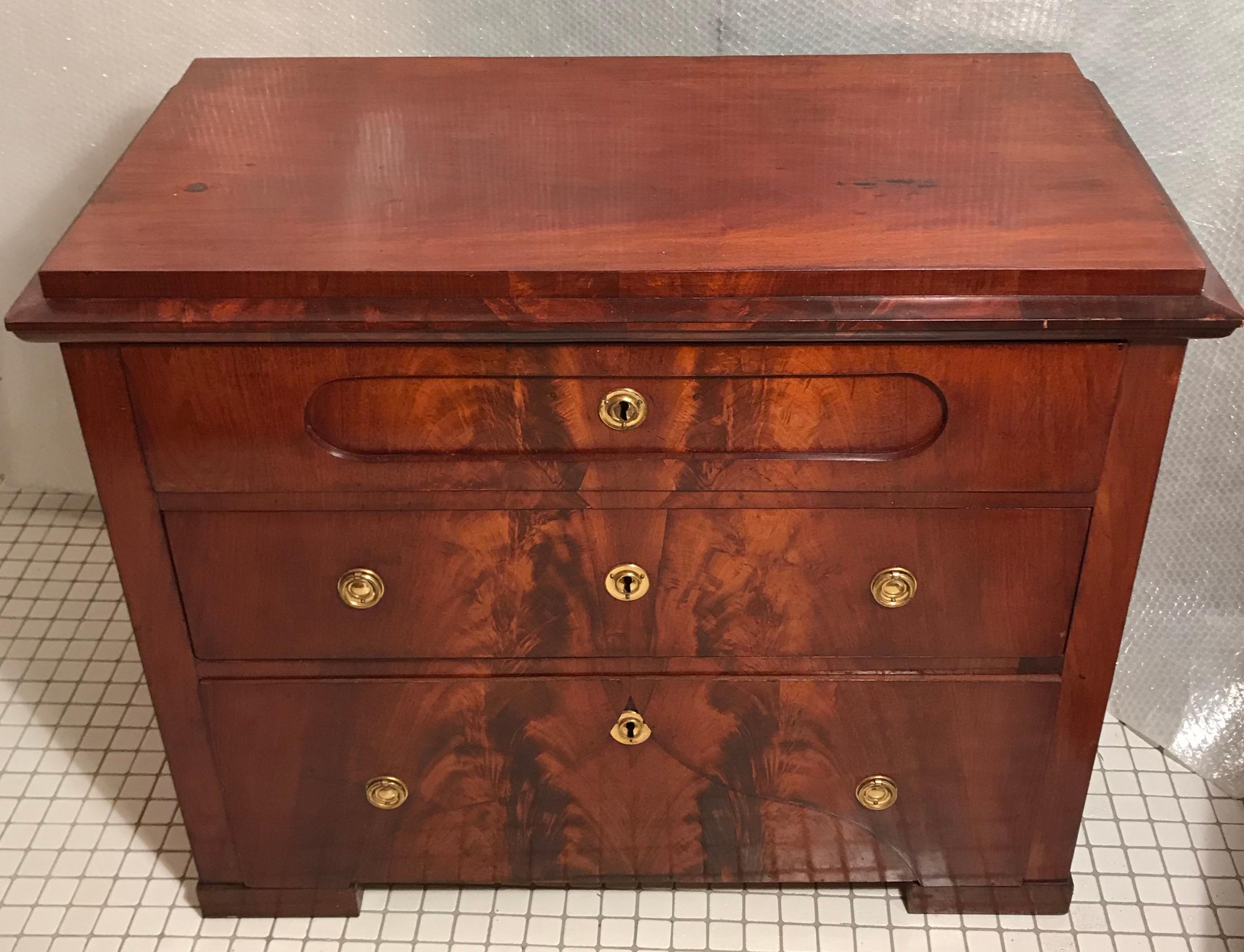 La cassettiera Biedermeier, che si distingue per la sua splendida impiallacciatura di mogano a specchio, è stata realizzata in Danimarca intorno al 1820. Ha tre cassetti e accessori originali in ottone. È in ottime condizioni, rifinito e lucidato