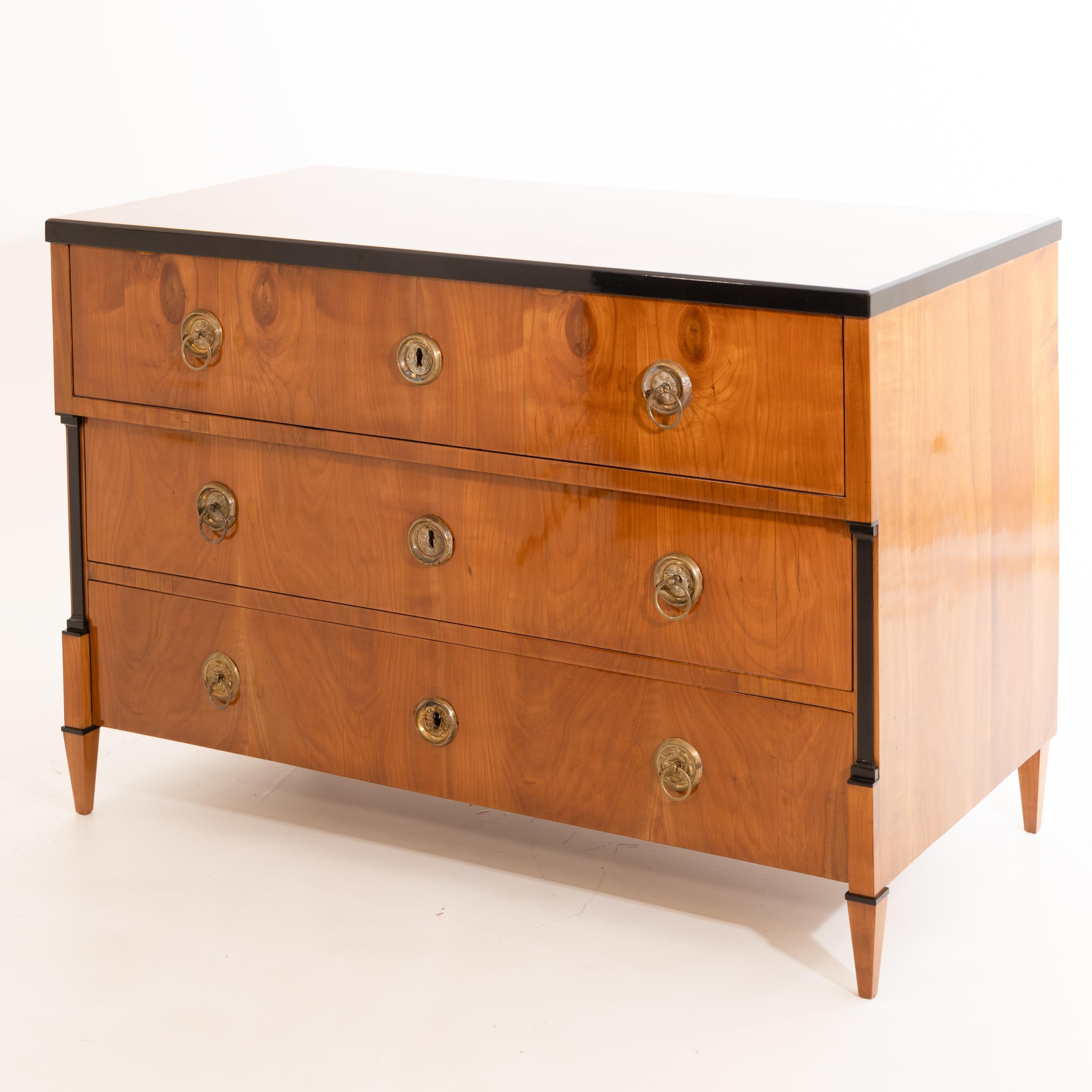 Three-drawer Biedermeier chest of drawers on square pointed feet with ebonised pilasters and an ebonised edge. The top drawer protrudes slightly and rests on the pilasters. The commode is veneered in cherry and decorated with disc-shaped bronze