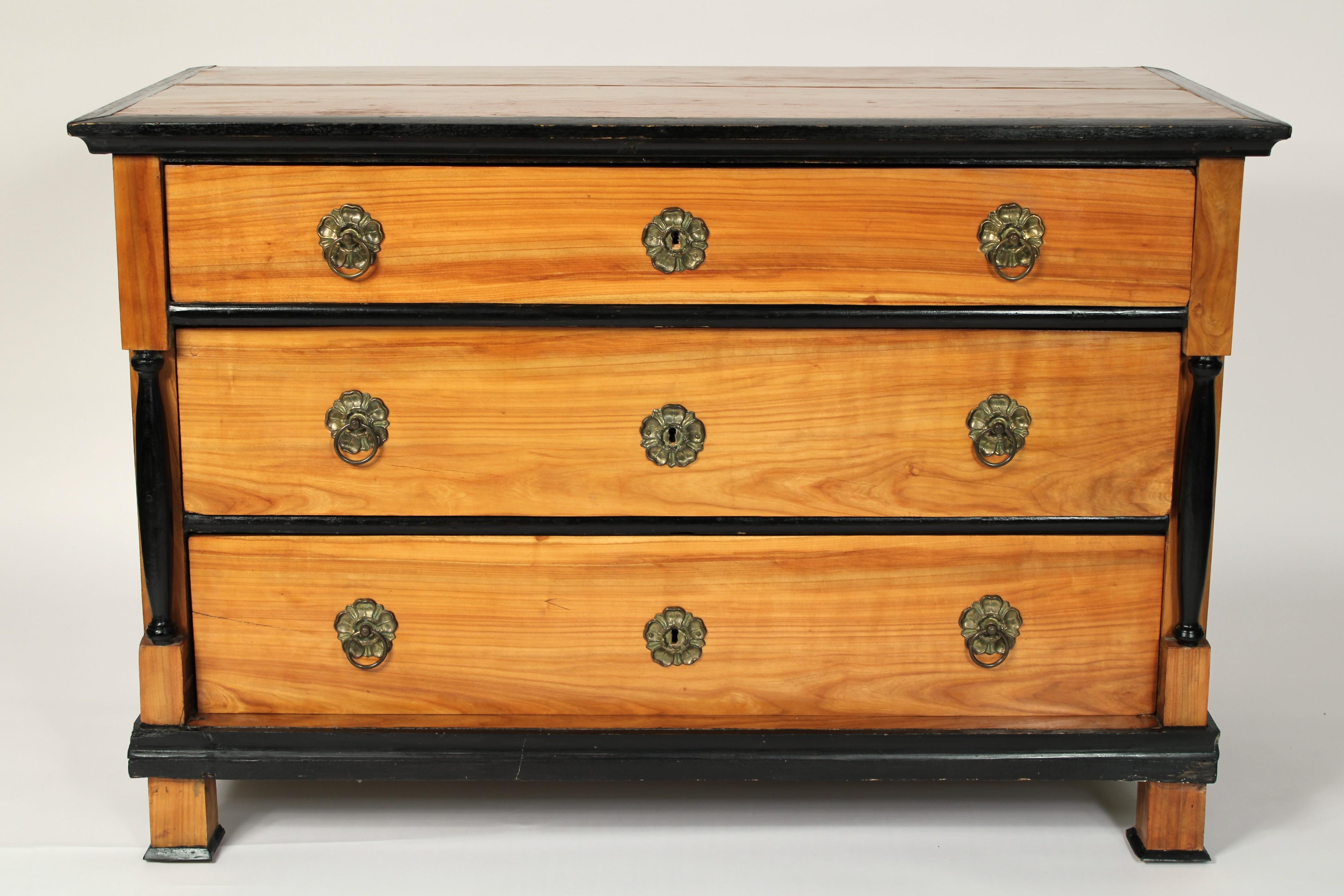 Biedermeier / Empire birch chest of drawers, circa 1830. With a rectangular birch top with a slightly chamfered ebonized front and side borders, 3 graduated drawers with later brass hardware, flanked by ebonized columns, resting on square feet.