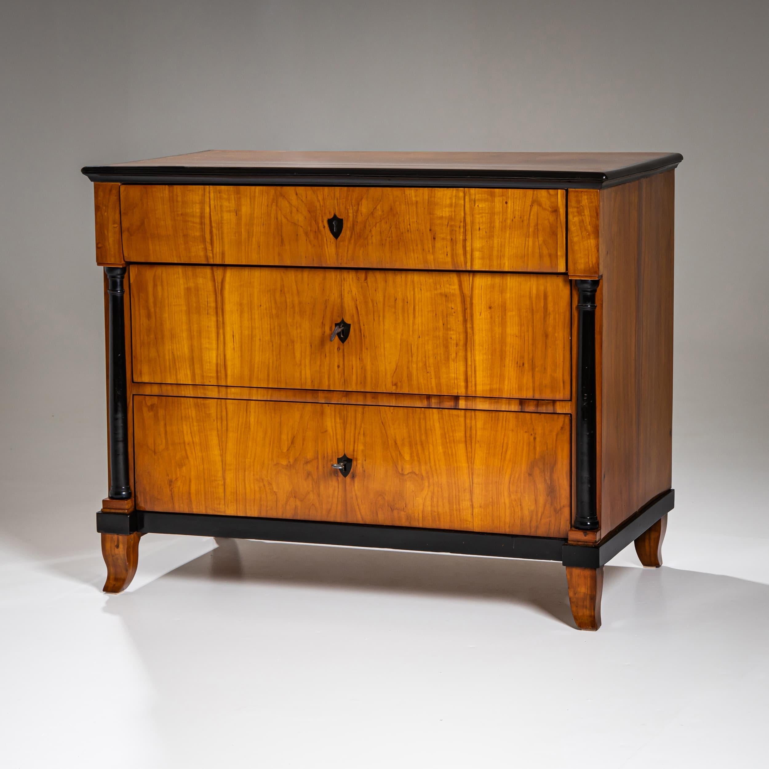 Elegante commode Biedermeier en merisier à trois tiroirs. La façade est flanquée de pilastres à demi-colonnes ébonisées, complétés par des écussons ébonisés et des accents à bords noirs. Posée sur des pieds légèrement incurvés, cette pièce a été