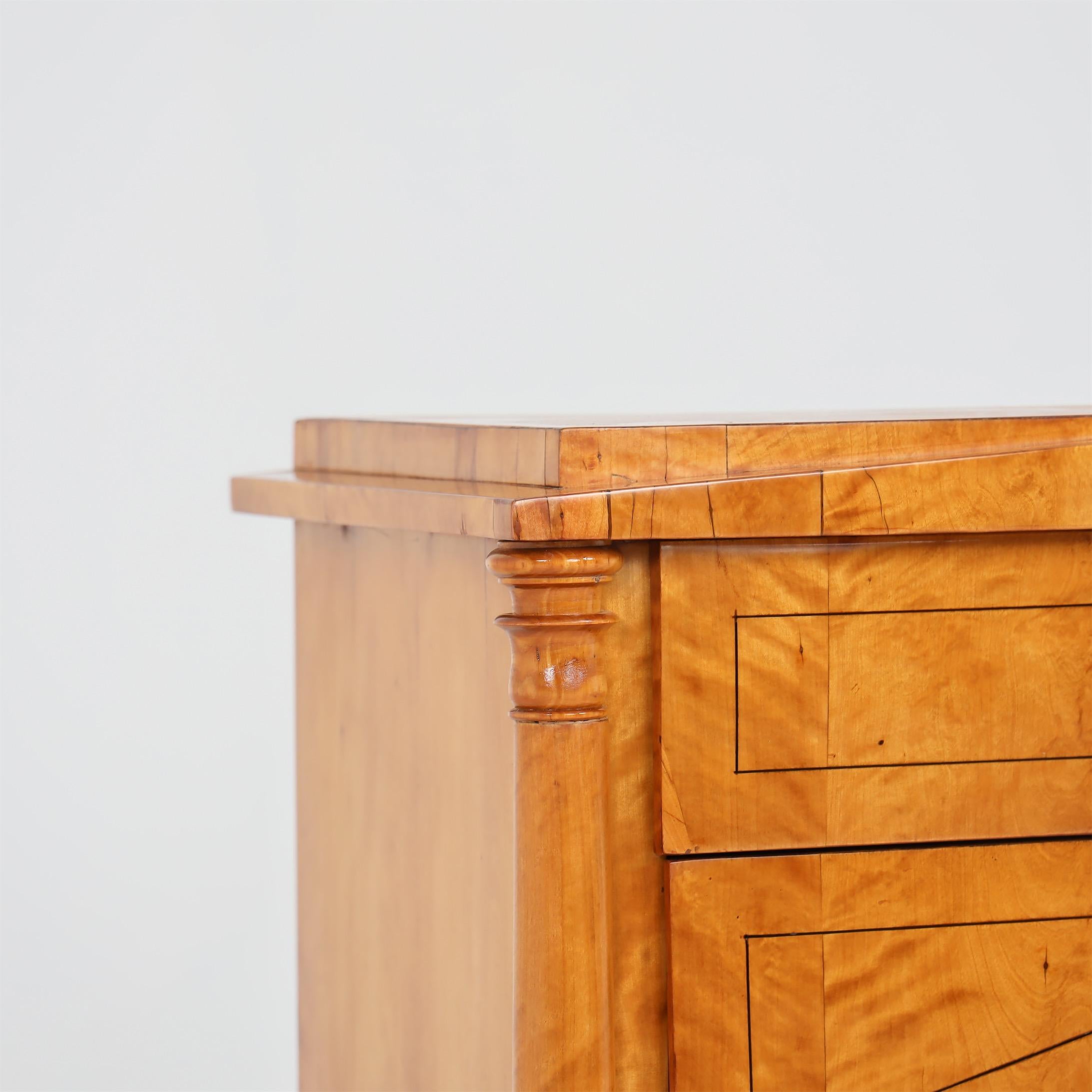 Biedermeier chest of drawers with three drawers standing on square legs. The body is veneered in birch and flanked by three-quarter columns at the corners, which support the surrounding edge executed as a pediment. The front shows thread inlays as
