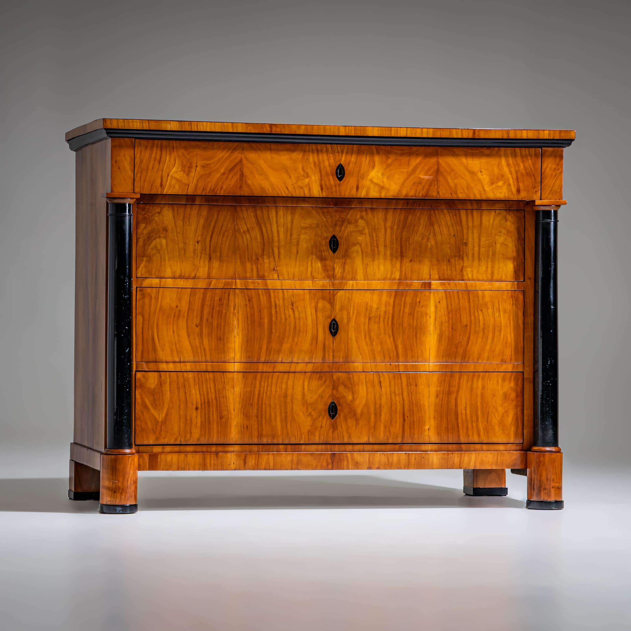 Four-drawer Biedermeier chest of drawers veneered in cherrywood, featuring ebonized full columns and key escutcheons. The piece rests on cylindrical front legs and square back legs. The top is framed with an ebonized molding. The slightly narrower