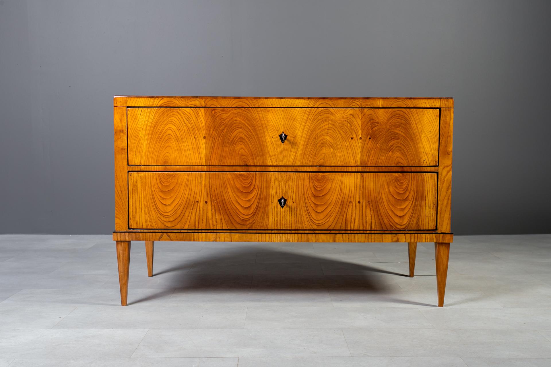 A beautifully proportioned 19th-century Austrian chest of drawers, veneered with Elm, professionally restored.
The chest stands on refined tapered legs, giving it a light and timeless silhouette.
A delicate black line inlay runs beneath the top and