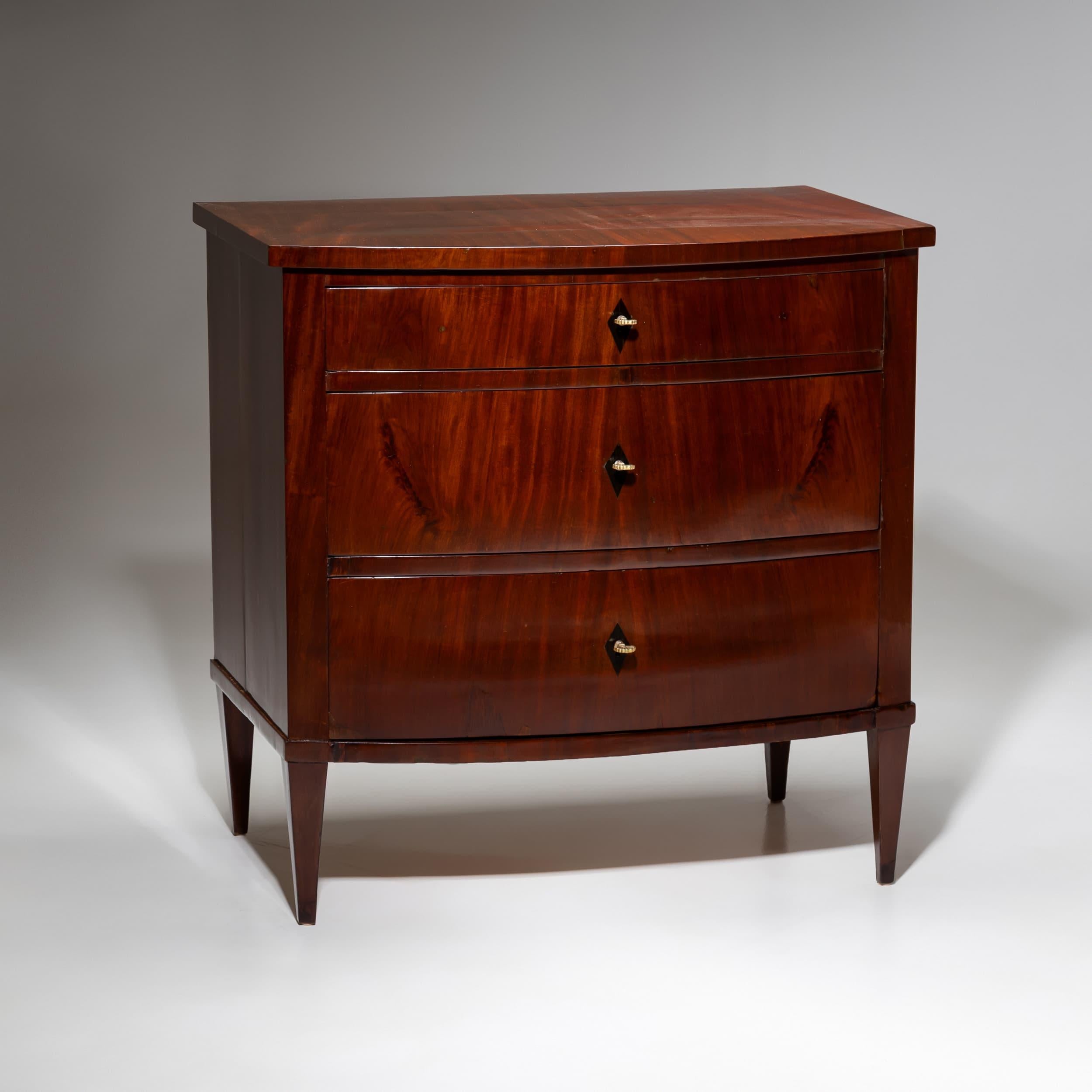 Commodo Biedermeier a tre cassetti su gambe a sezione quadrata. La parte anteriore leggermente convessa conferisce al pezzo una silhouette raffinata. Impiallacciata in mogano, con scudi a chiave disegnati come intarsi a forma di losanga ebanizzata.