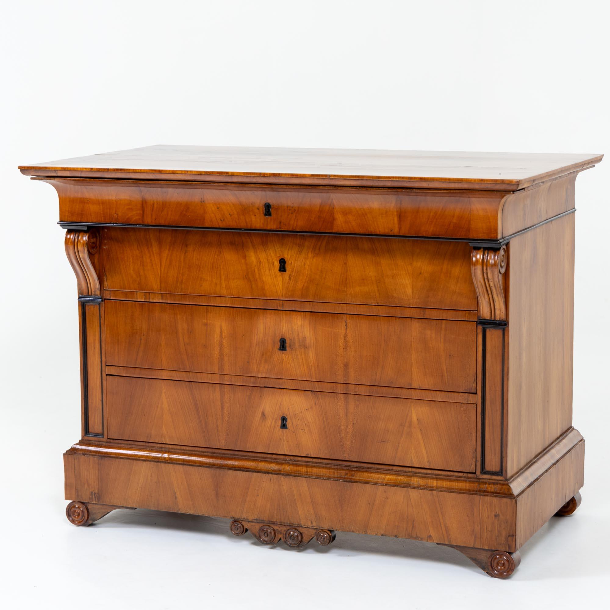 Biedermeier chest with volute legs, veneered in cherry with four drawers. The corpus with flanking volute capitals and ebonised mouldings rises above the wide plinth zone. The head drawer is slightly lower and is finished off by the profiled top.
