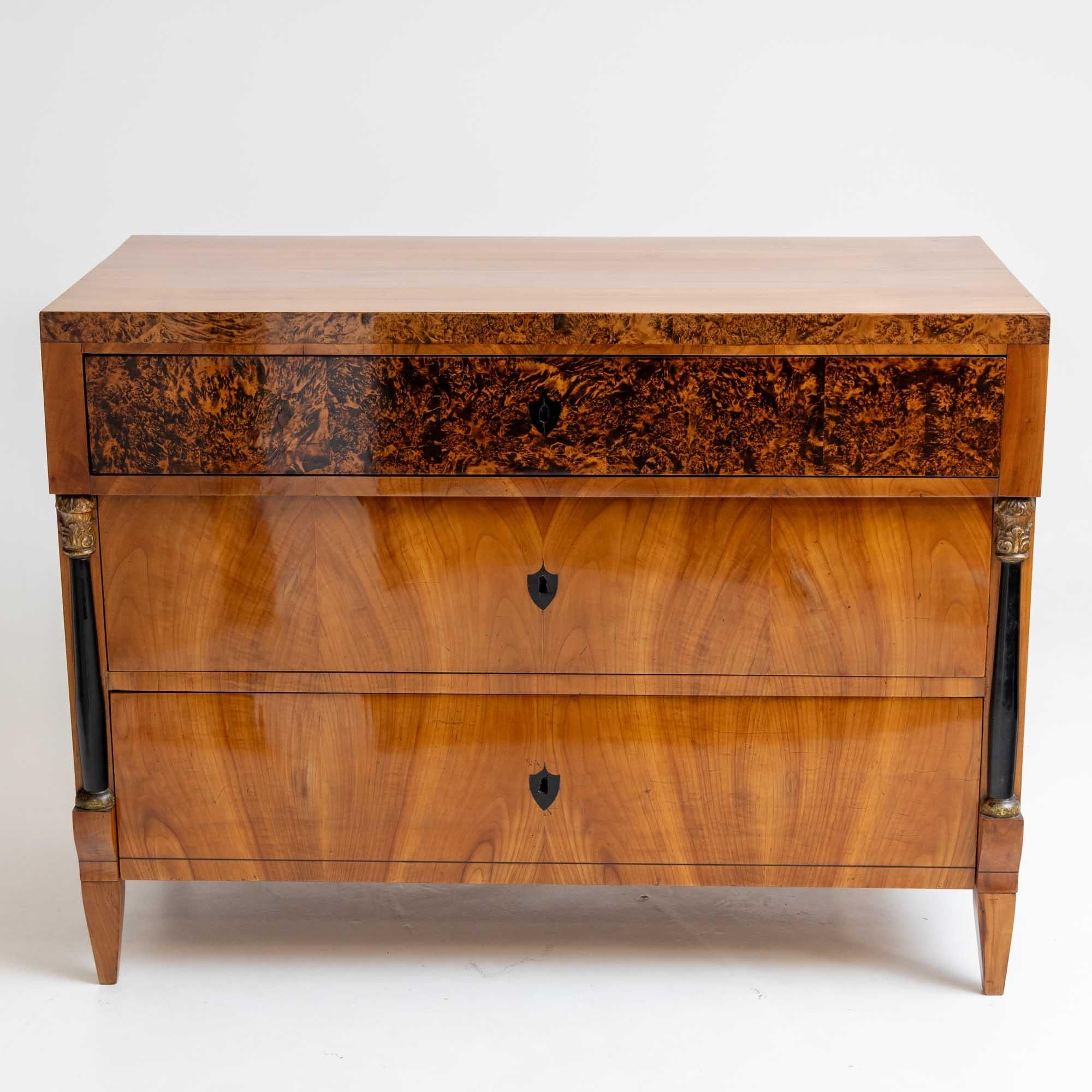 Biedermeier chest of drawers crafted from cherry wood and adorned with burl inlays. The key plates take the form of ebonized shields. Ebonized half-columns with gold-patinated capitals and bases flank the two lower drawers, adding a touch of