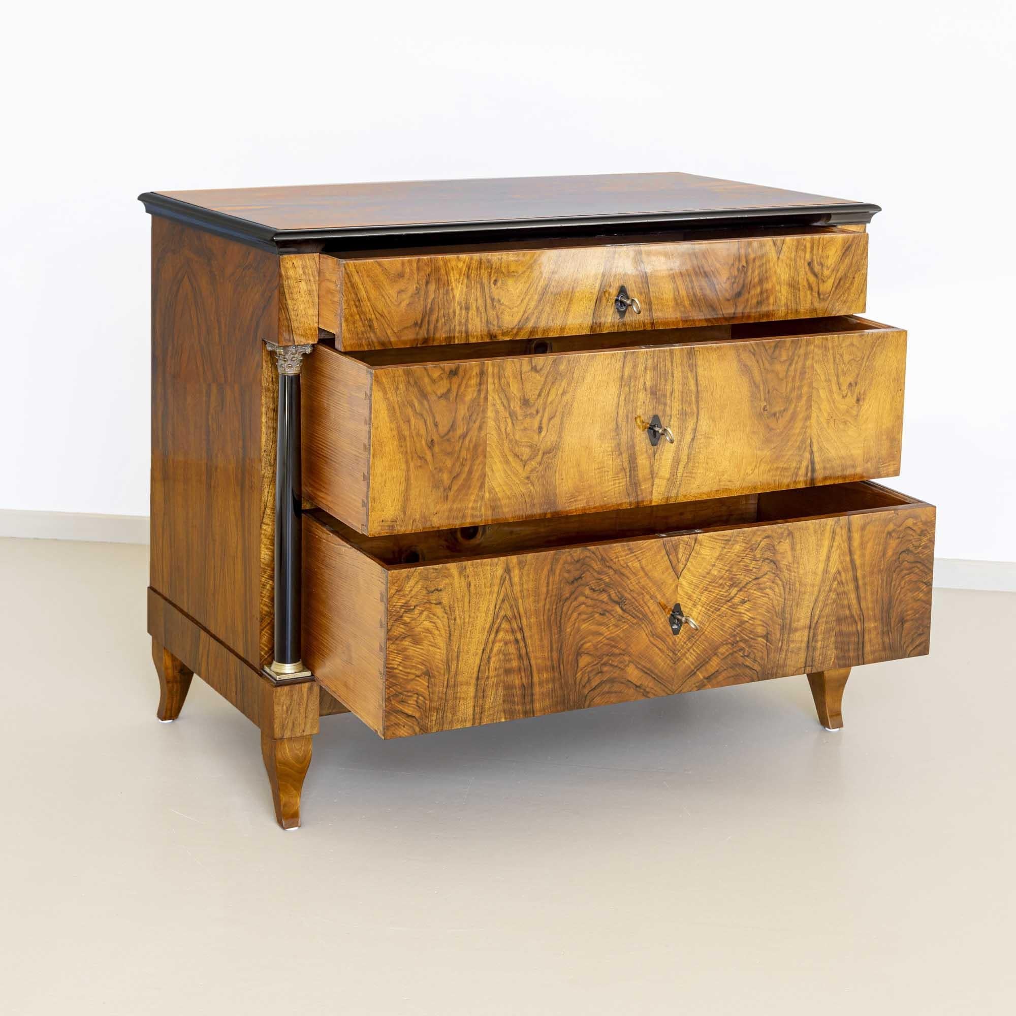 Biedermeier chest of drawers on S-shaped legs with ebonized columns, bases and brass capitals. The surrounding profiled edge is also ebonized. The chest of drawers offers plenty of storage space with three drawers. The chest of drawers is veneered