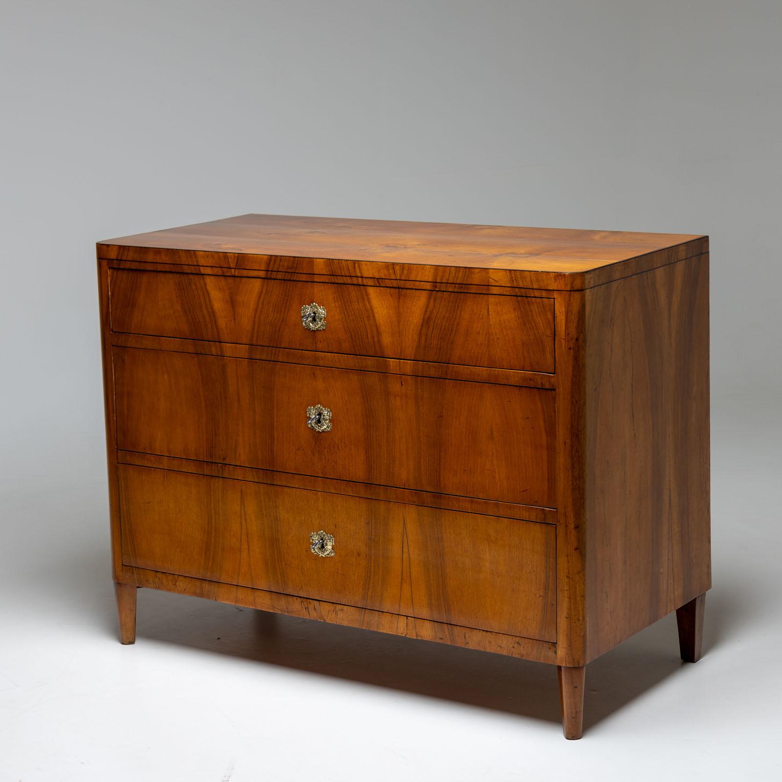 Cassettiera Biedermeier con tre cassetti impreziositi da accessori in bronzo dorato a fuoco che fungono da placchette per le chiavi. Poggiata su gambe affusolate, questa cassapanca è impiallacciata in noce. Restaurata professionalmente e lucidata a