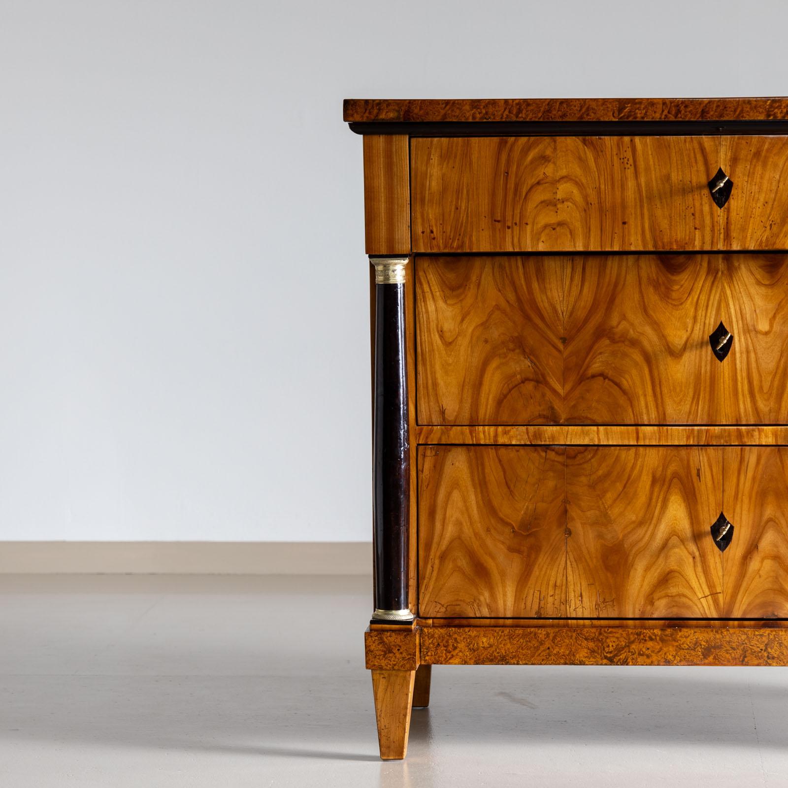 Three-drawer Biedermeier chest of drawers with flanking columns with brass bases and capitals. The chest of drawers is veneered in cherry and thuja burl. The key plates are ebonized. The chest of drawers has been polished and restored by hand. 