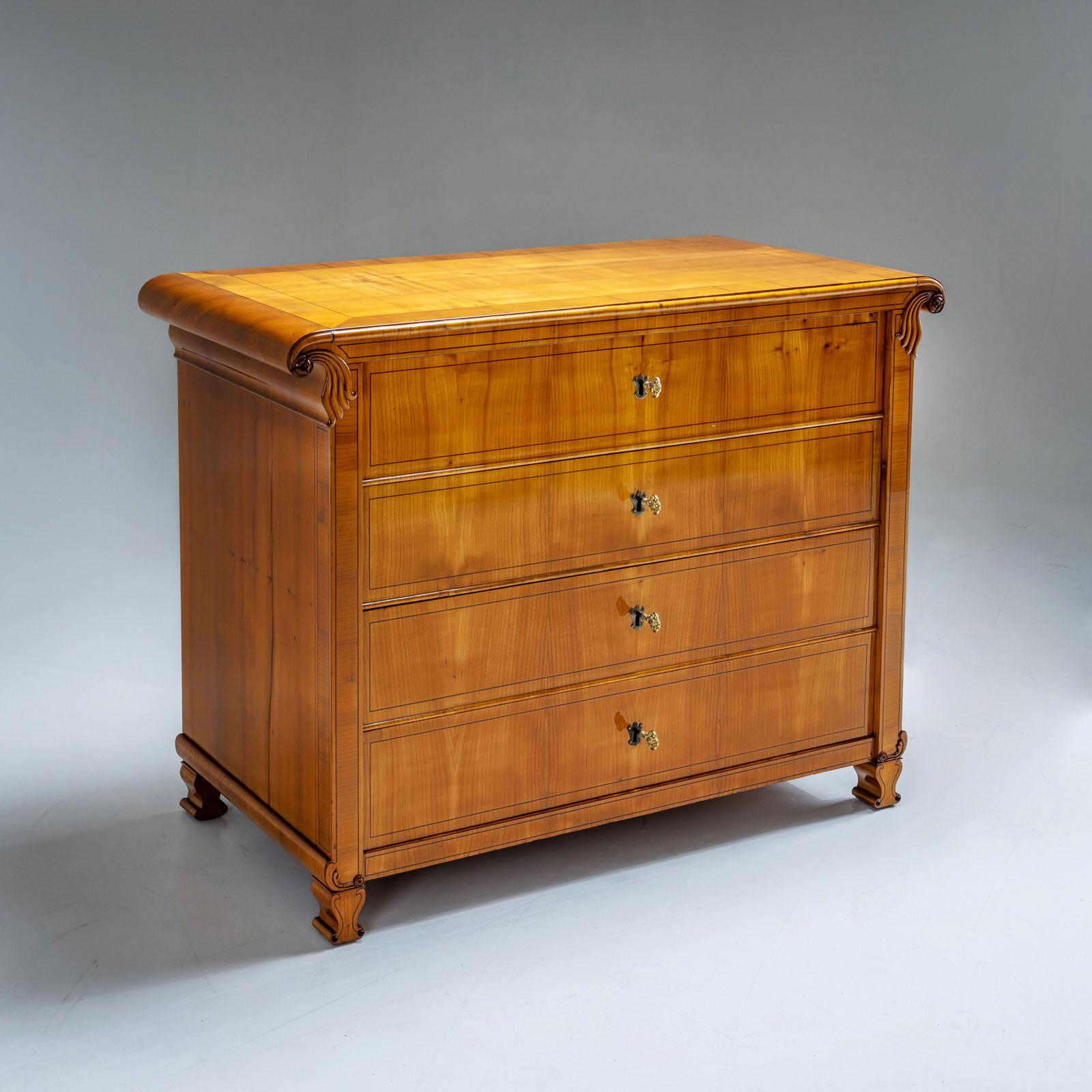 Cassettiera Biedermeier con quattro cassetti e un piano superiore che forma volute e piedi a forma di vaso. La cassettiera è impiallacciata in ciliegio. Le traverse sono profilate e le superfici sono incorniciate da intarsi di fili. La cassettiera è