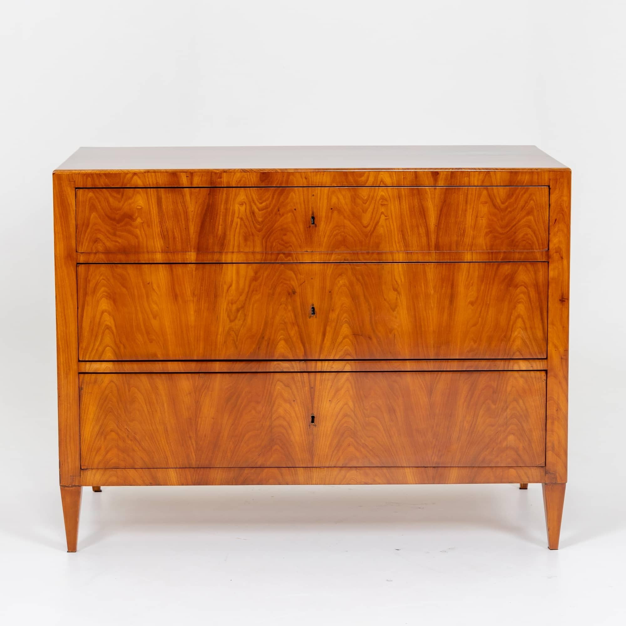 Biedermeier chest of drawers on square tapered legs with a straight-lined body and three drawers. The chest of drawers is veneered in cherry and has been polished by hand. The top drawer is slightly narrower than the bottom two. The chest of drawers
