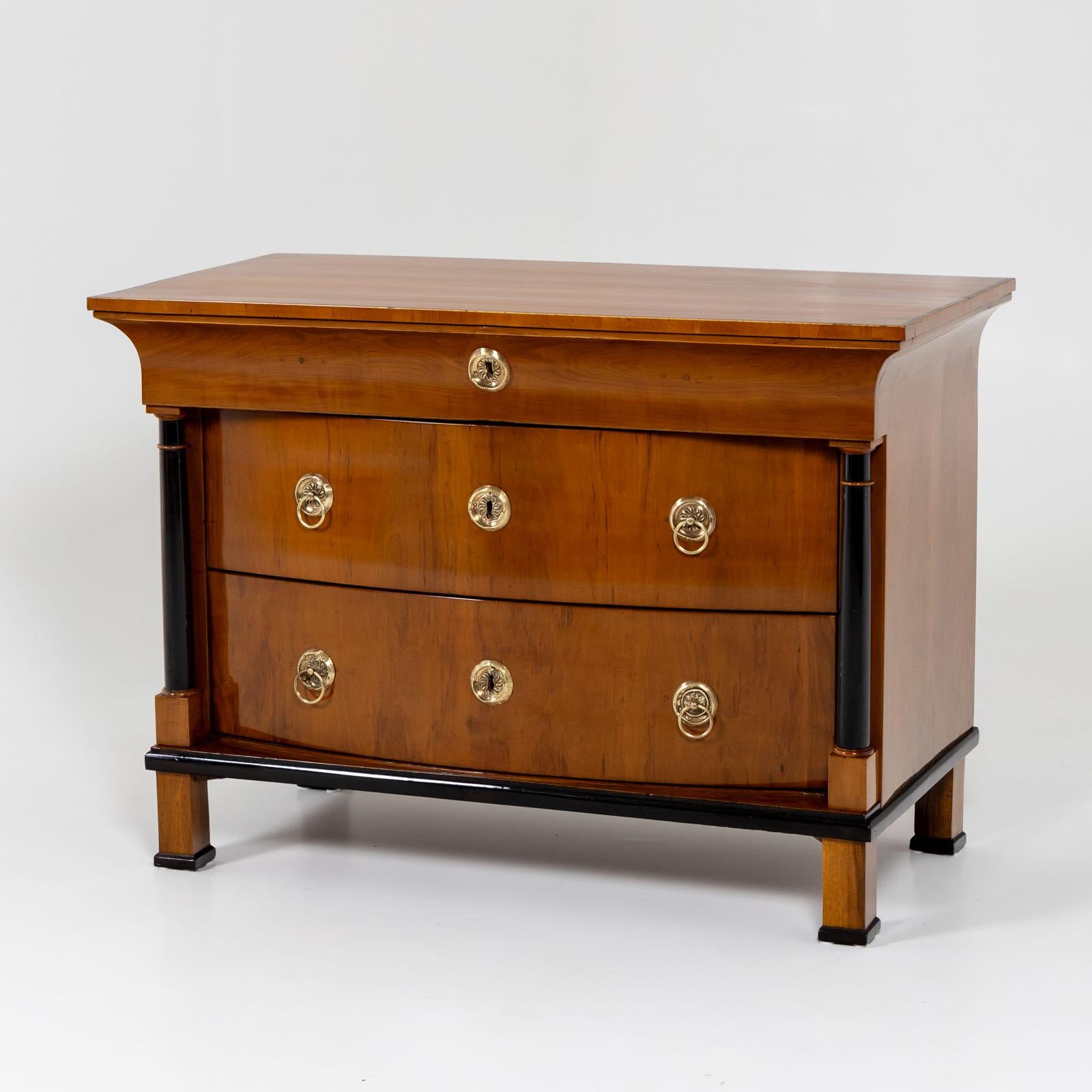 Biedermeier chest of drawers with cambered front and three drawers. The two lower drawers are flanked by ebonized columns. The chest of drawers stands on square feet and is veneered in pear wood. Disc-shaped brass fittings adorn the front. The head