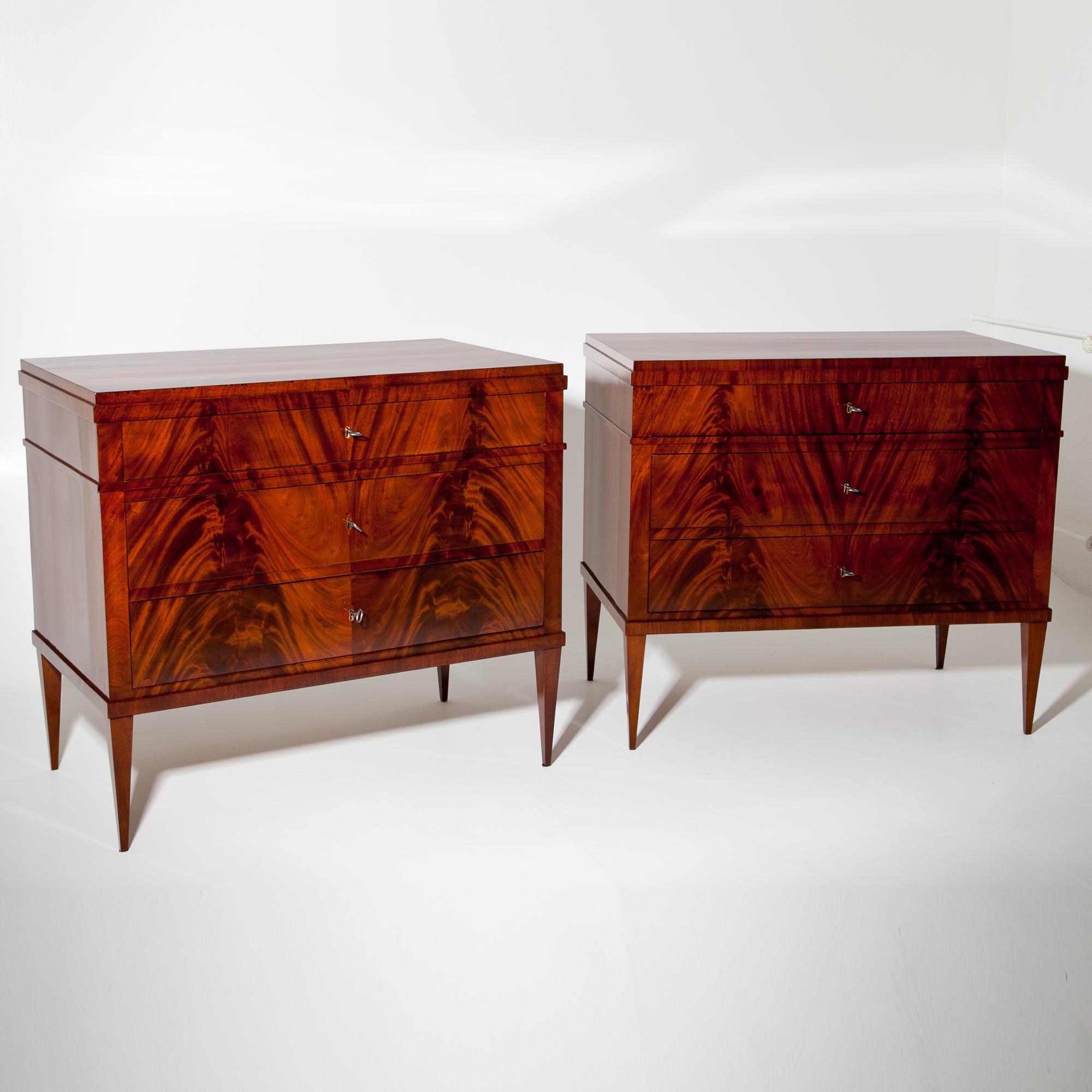 Pair of mahogany-veneered chests of drawers, standing on slender tapered legs. Profiled trusses accentuate the top drawer. The front shows a beautiful flamed mahogany veneer. The commodes were professionally refurbished and hand-polished.