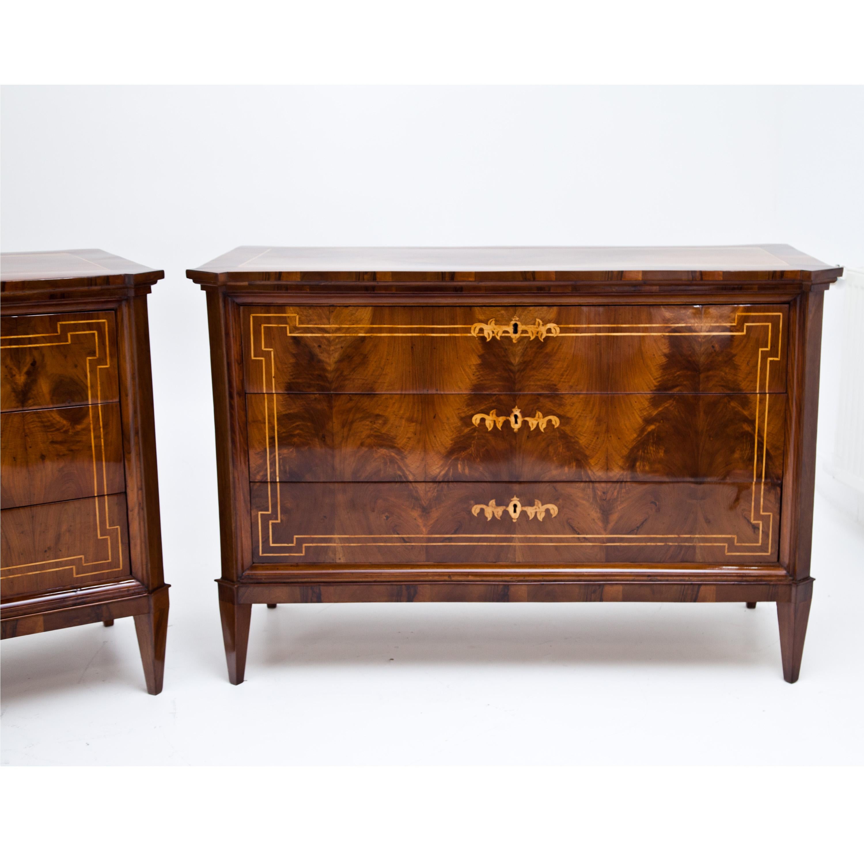 Pair of Biedermeier chests of drawers with three drawers on square pointed feet. The body with bevelled corners and profiled strips. The drawers and top shelf are framed with thread inlays. The keyholes are decorated with additional inlays in the
