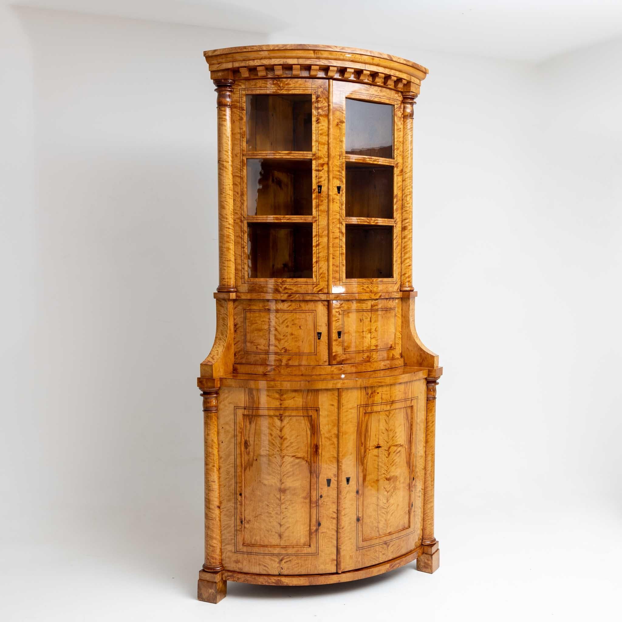 A Biedermeier corner display case with a curved front, a two-door lower section with flanking column decoration, a central section also with two doors and a display case top with glazed doors and three shelves. The corner display case is veneered in