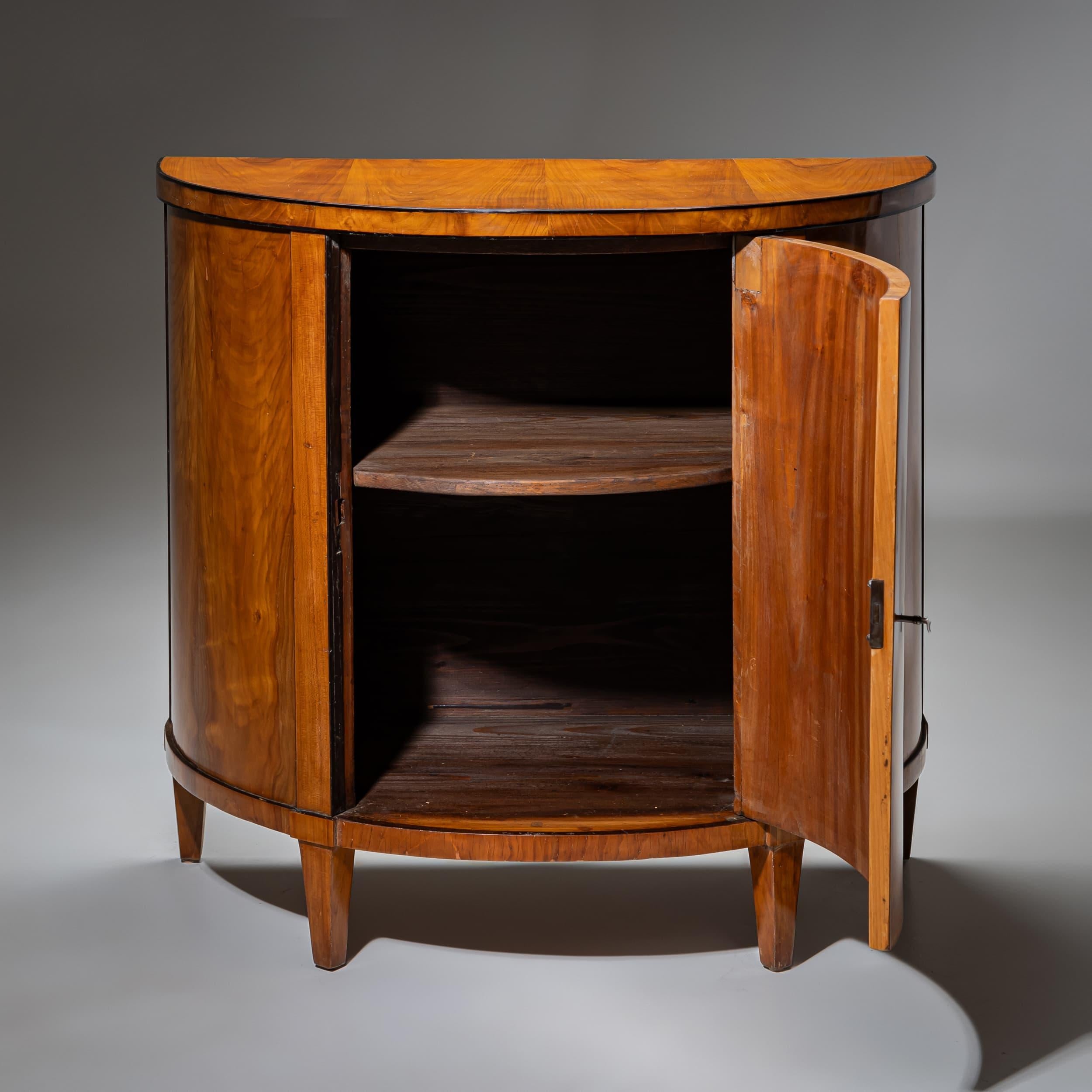 Ein eleganter Biedermeier-Schrank, der um 1820 in Süddeutschland gefertigt wurde. Der Schrank steht auf schlanken, spitz zulaufenden Beinen mit dunklen Akzenten, die einen raffinierten Kontrast zum warmen Kirschholz bilden. Die Vorderseite ist mit