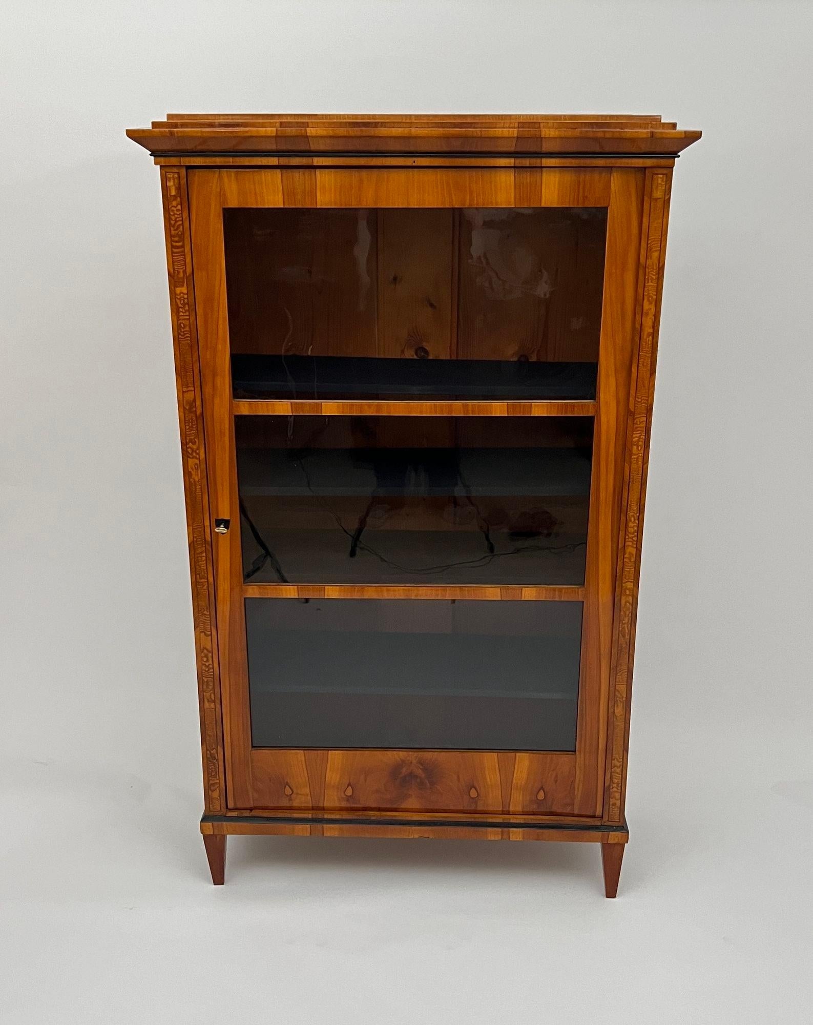 Elegante vitrine ou bibliothèque Biedermeier du sud de l'Allemagne vers 1820.

Bois de cerisier clair plaqué sur bois tendre. Partiellement ébonisé.
Placage de ronce de frêne incrusté sur les pilastres latéraux et encadré par une incrustation de