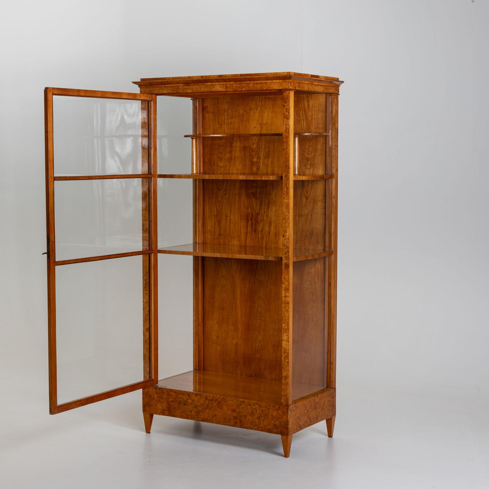 Single-door Biedermeier display cabinet with a three-sided glazed front and two shelves as well as a small curved shelf supported by small columns. The display cabinet is veneered in thuja root and cherry. It has been restored and polished by hand.