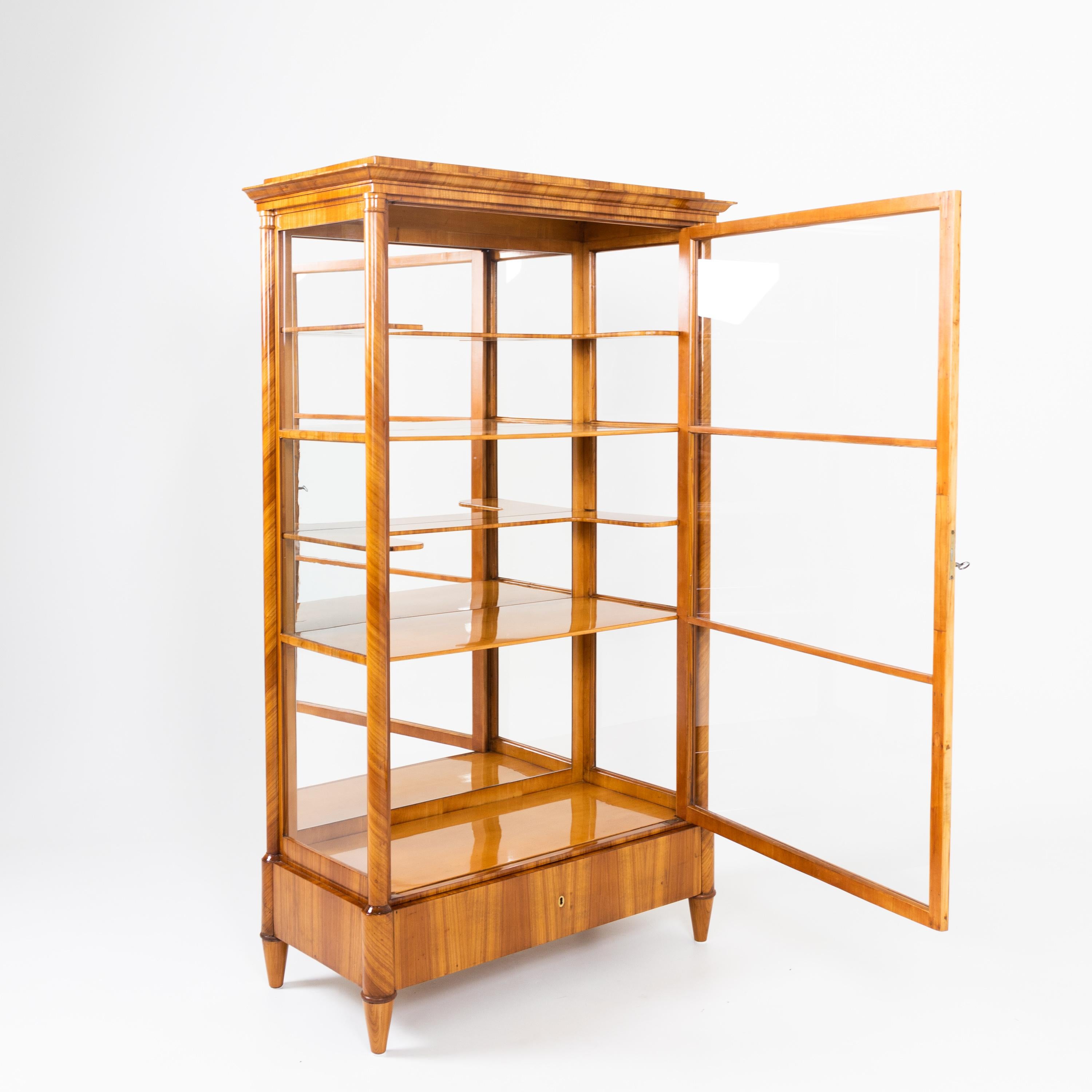 Three-sided glazed display case with one drawer standing on conical feet. The corners of the corpus are flanked by columns of solid cherry and support the multiple profiled cornice. The interior with mirrored back panel consists of four shelves, two