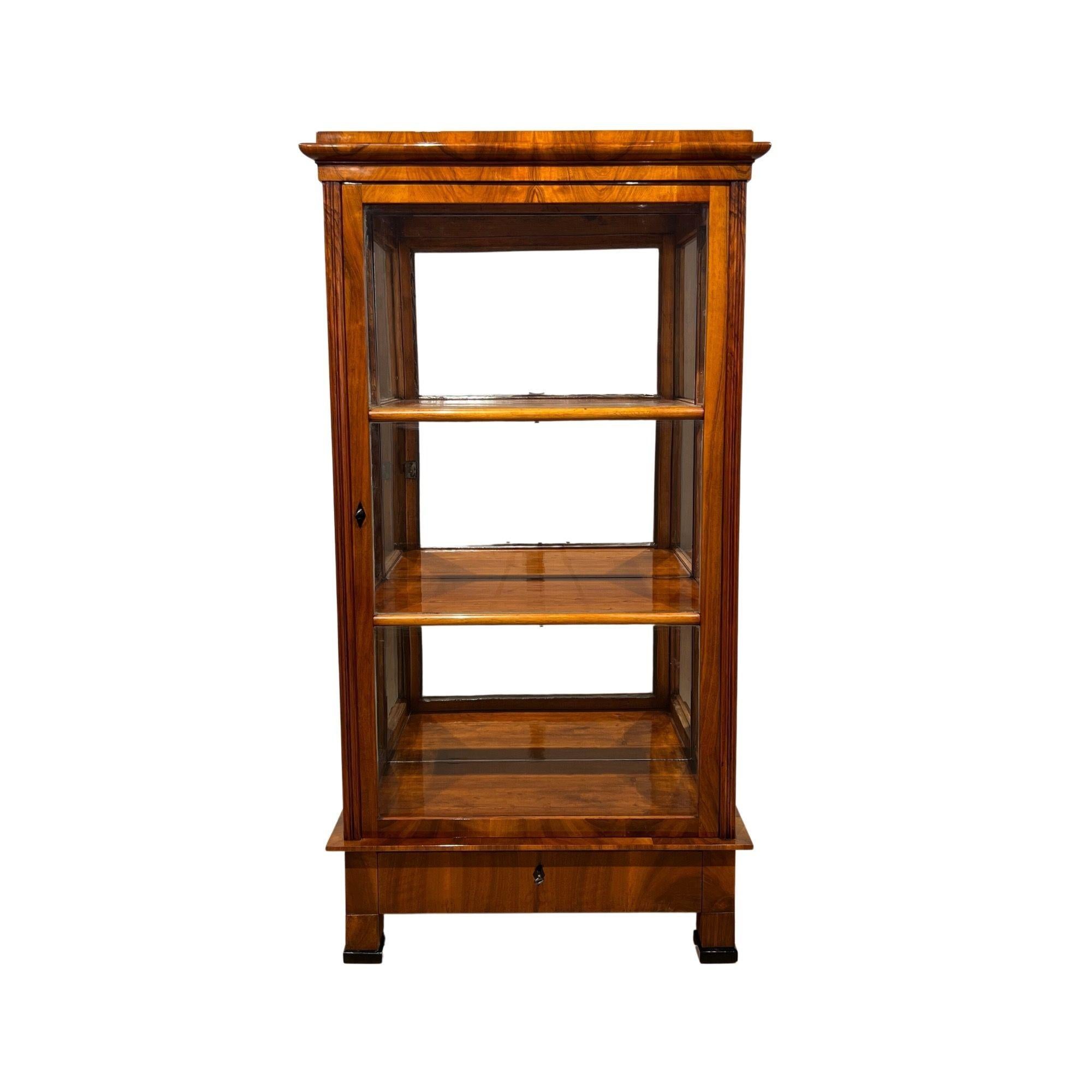 Beautif three-sided glazed Biedermeier display case, vitrine or showcase in walnut from south Germany around 1830. 
Walnut veneered on softwood. Classicist shaped cornice. Fluted side lisettes. Below one drawer with lock. Inside 2 shelves with 3
