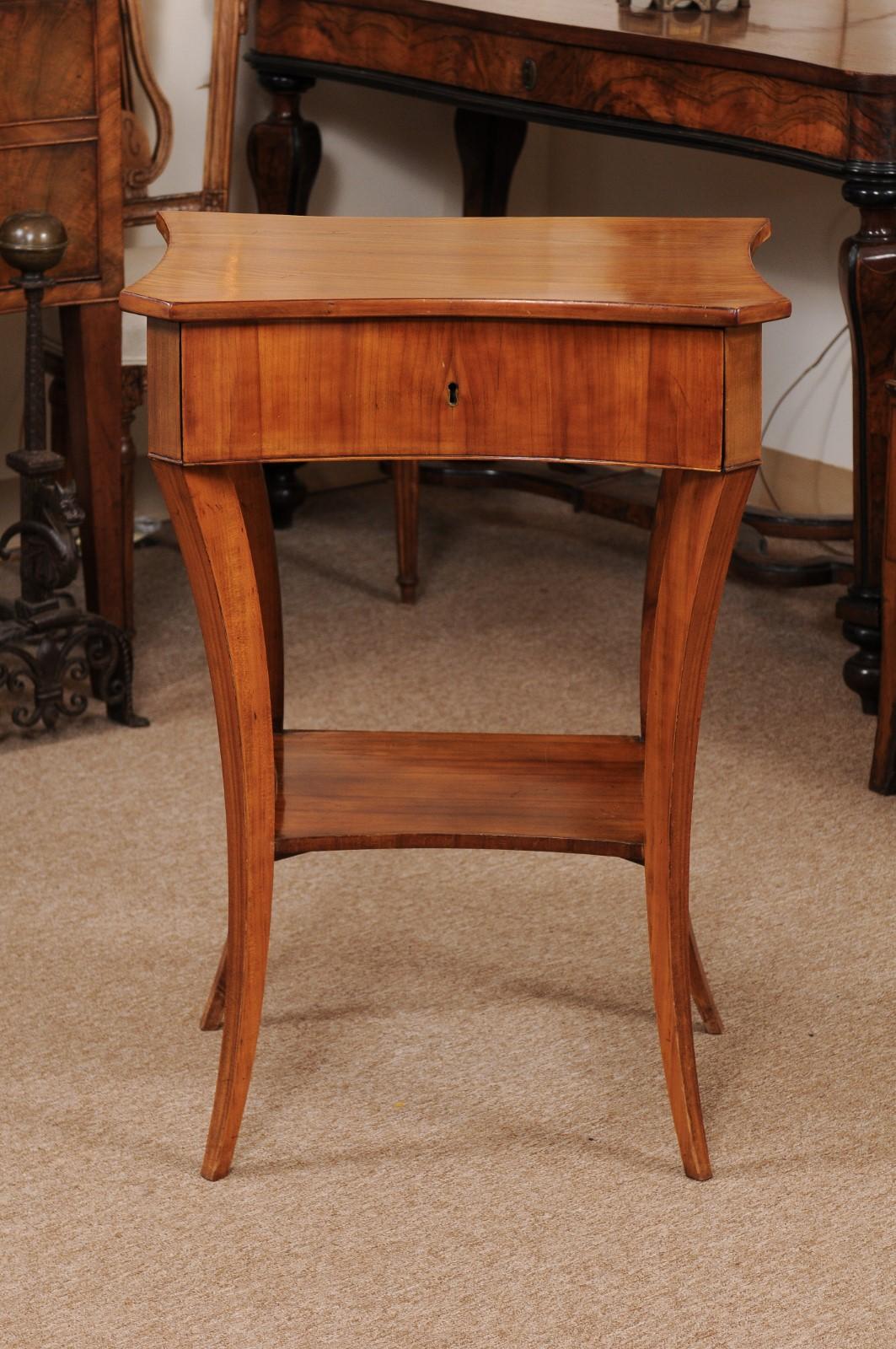 A Biedermeier early 19th century side table in ash and fruitwood with concave formed top, drawer in frieze and lower shelf joined by tapered splayed legs.