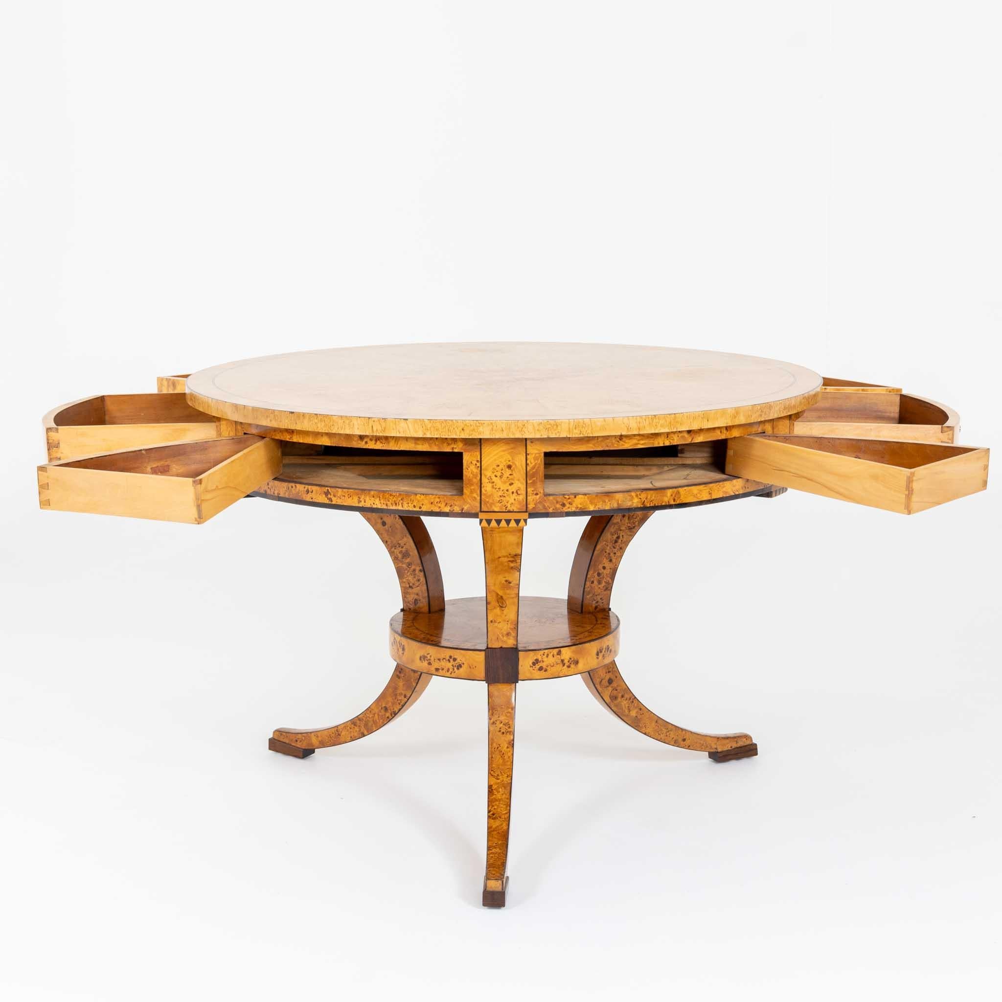 This exquisite game table features three curved legs with intermediate bracing and a deep straight apron adorned with indicated dentil strip. It boasts six drawers and a radially veneered round tabletop. The light birch veneer is highlighted at the