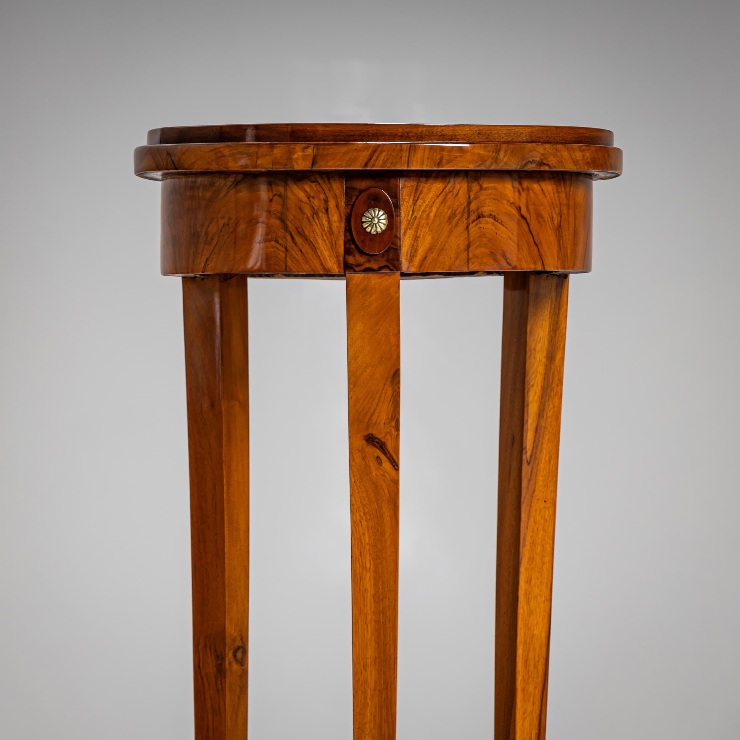 Elegant guéridon Biedermeier reposant sur trois pieds effilés et légèrement incurvés, reliés par une entretoise trilobée ornée d'un motif d'urne. Le tablier lisse est rehaussé de petites rosettes en laiton. Le plateau rond est encadré par un bord