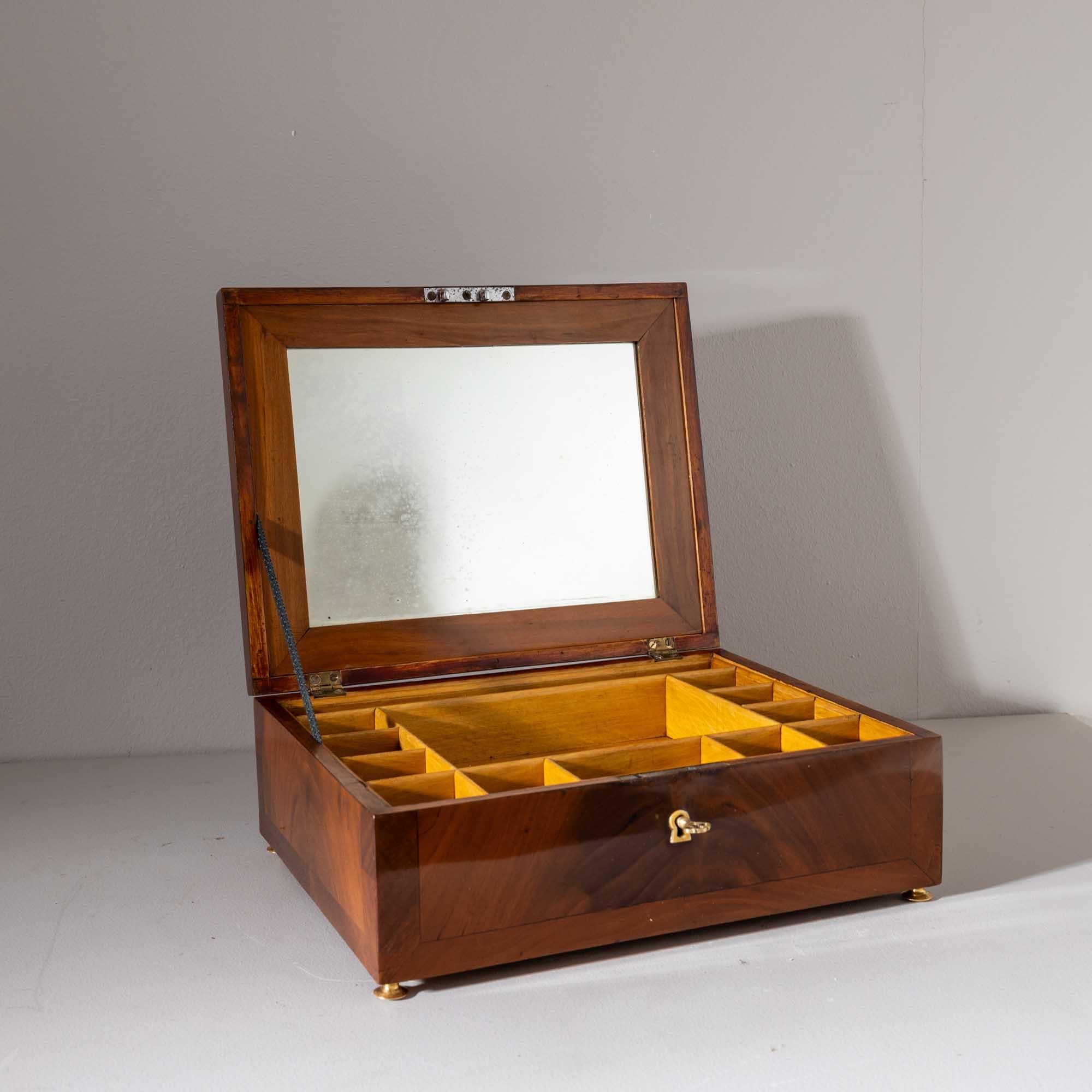 Small rectangular casket with slightly beveled lid and filling-shaped veneer pattern made of walnut. The casket stands on small brass feet. Inside it is mirrored and equipped with small compartments.