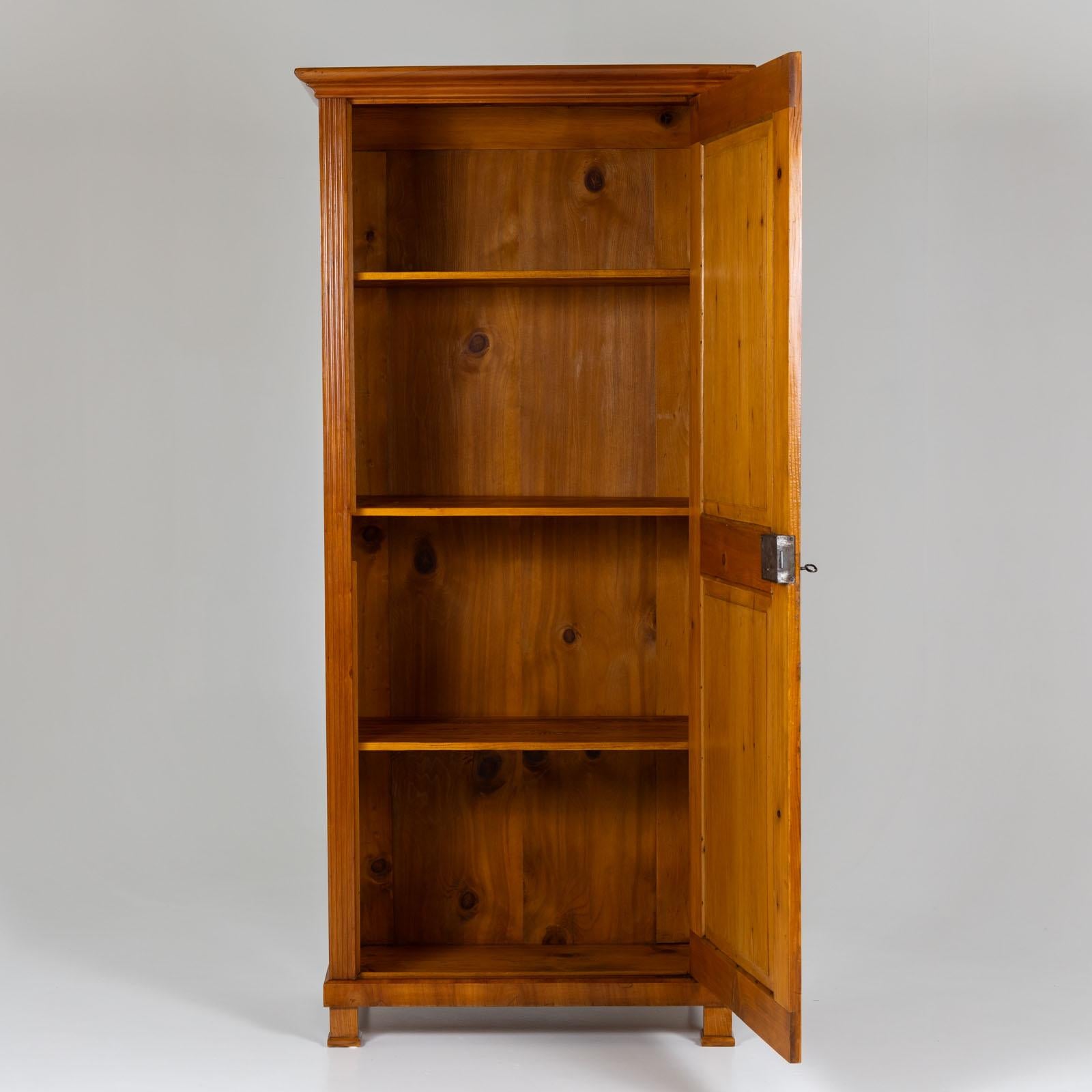 Biedermeier linen cupboard in cherry veneer with one door and square feet. The door is divided into two panels and the pilaster strips are fluted. The inside of the cupboard is fitted with three shelves and has been restored and polished by hand. 