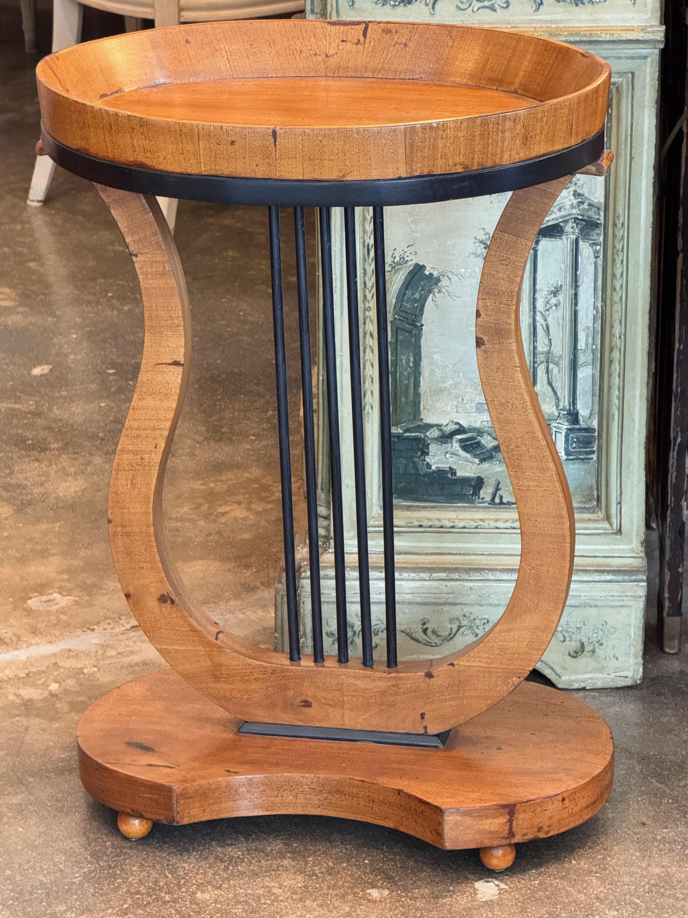 Petite table d'appoint de style Biedermeier avec une base en forme de lyre et un plateau ovale à bord relevé. Le cadre en bois aux tons chauds est complété par des accents peints en noir et des supports de tringles, le tout reposant sur des pieds en