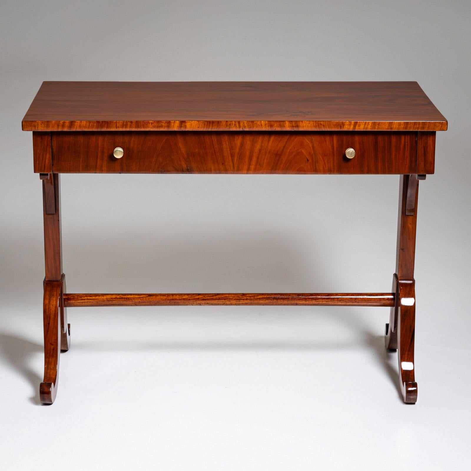 Un'elegante scrivania Biedermeier realizzata in mogano. Il pezzo è caratterizzato da eleganti gambe biforcute collegate da una barella liscia. Il suo piano rettangolare comprende un ampio cassetto dotato di pomelli rotondi in ottone. La scrivania è