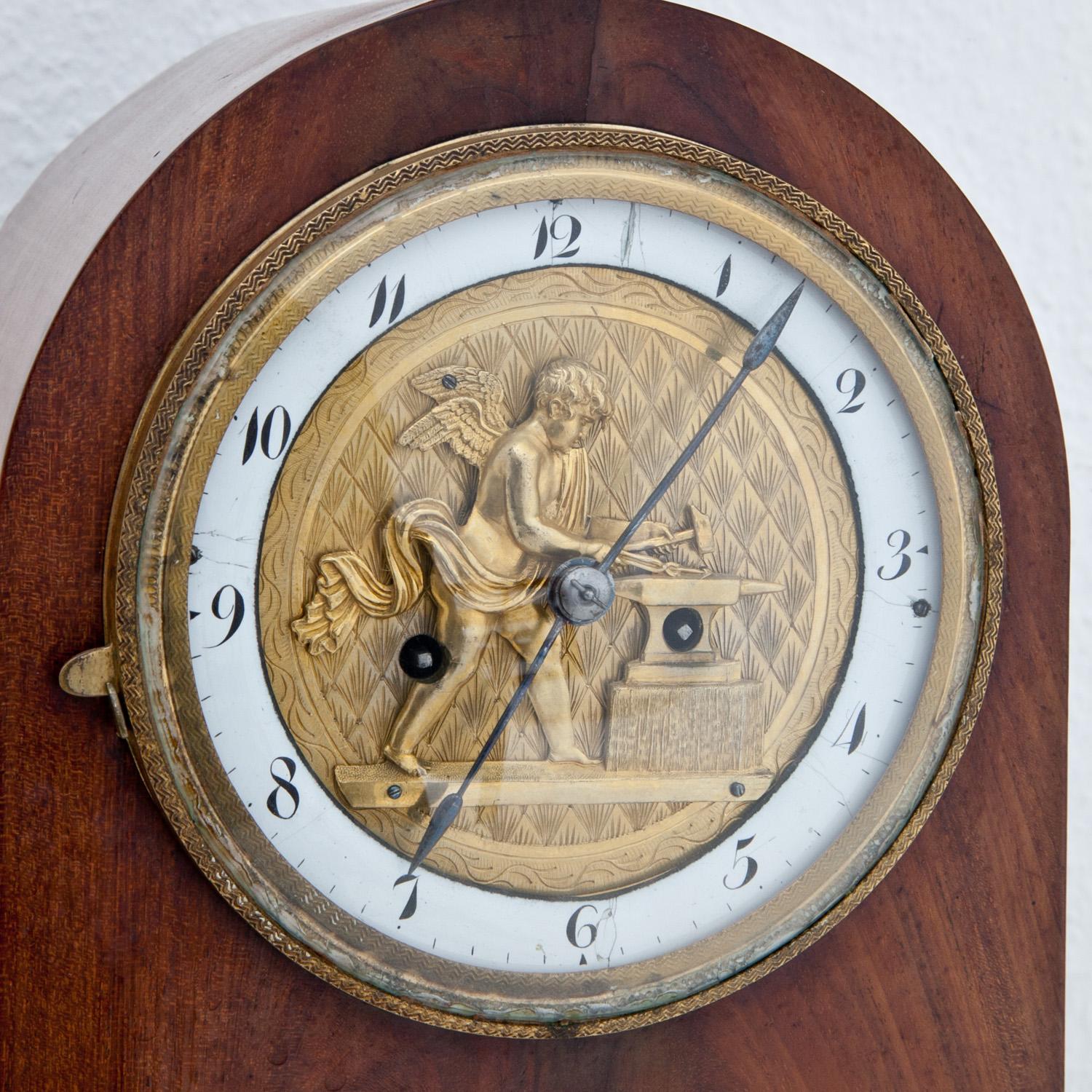 Biedermeier clock in a rounded mahogany case with an enamel ring with Arabic numerals and a bronze depiction of an angel with an anvil while forging. Underneath sun rays in a semi-circle on a black background.