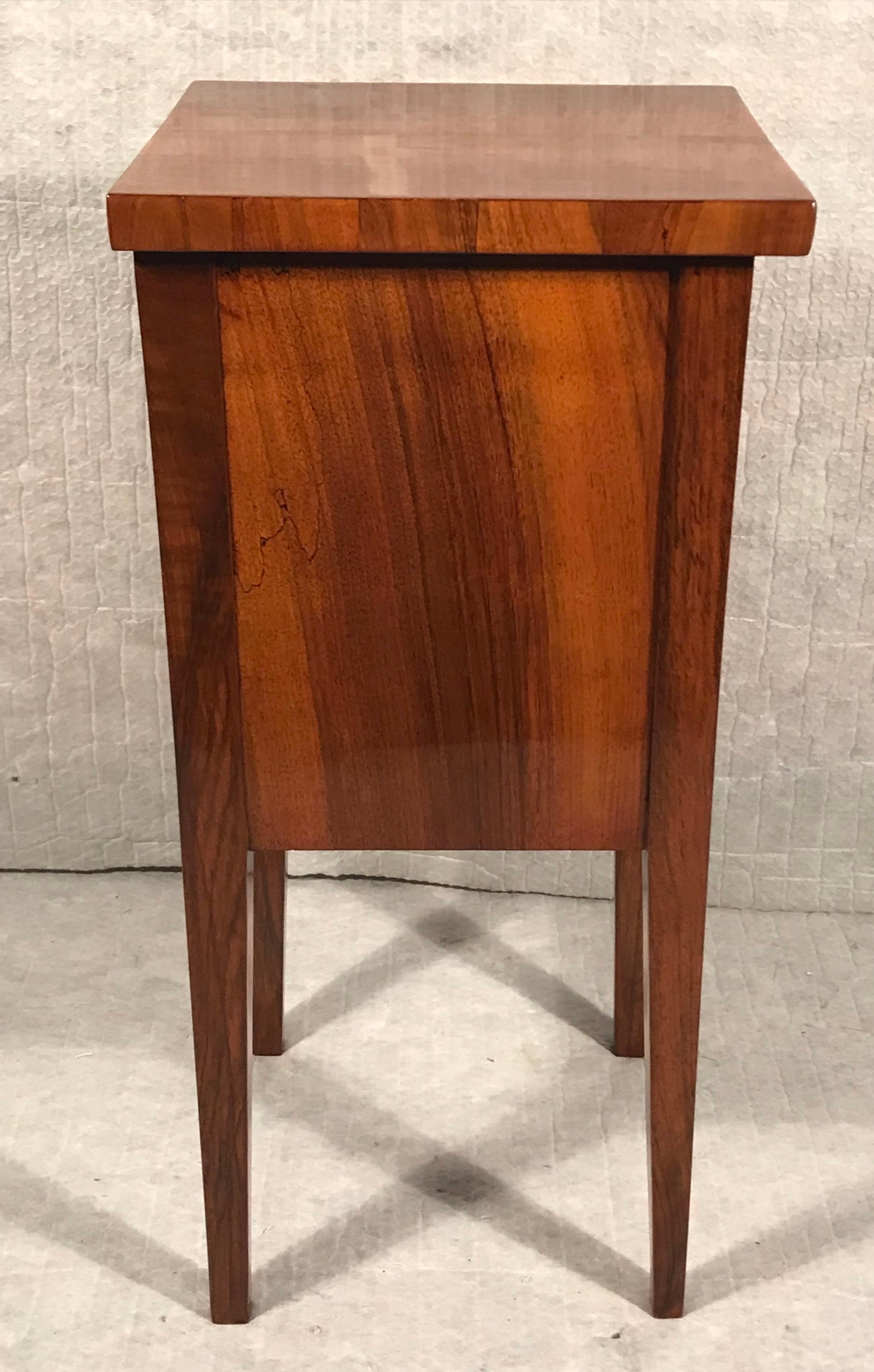 Descubre una exquisita mesilla de noche Biedermeier original del sur de Alemania, que data de 1820. Esta elegante pieza presenta un diseño recto adornado con una impresionante chapa de nogal en los cuatro lados. Apoyada en cuatro esbeltas patas, la