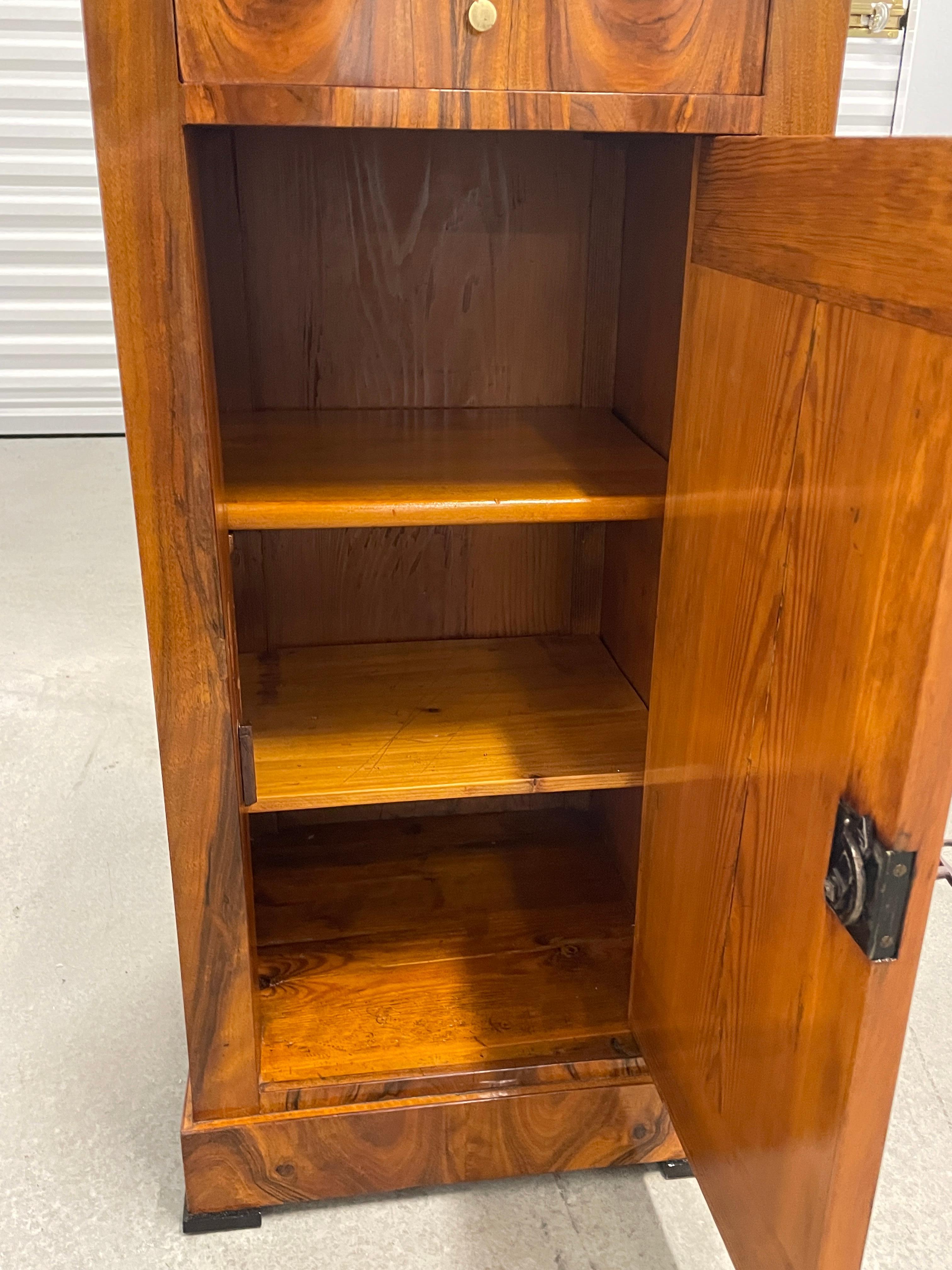 This original 19th century Biedermeier Nightstand has a very pretty walnut veneer. 
The small cabinet has one door, behind the door is a shelf. Above the door is a drawer. The Biedermeier Nightstand is in very good refinished condition. It has a
