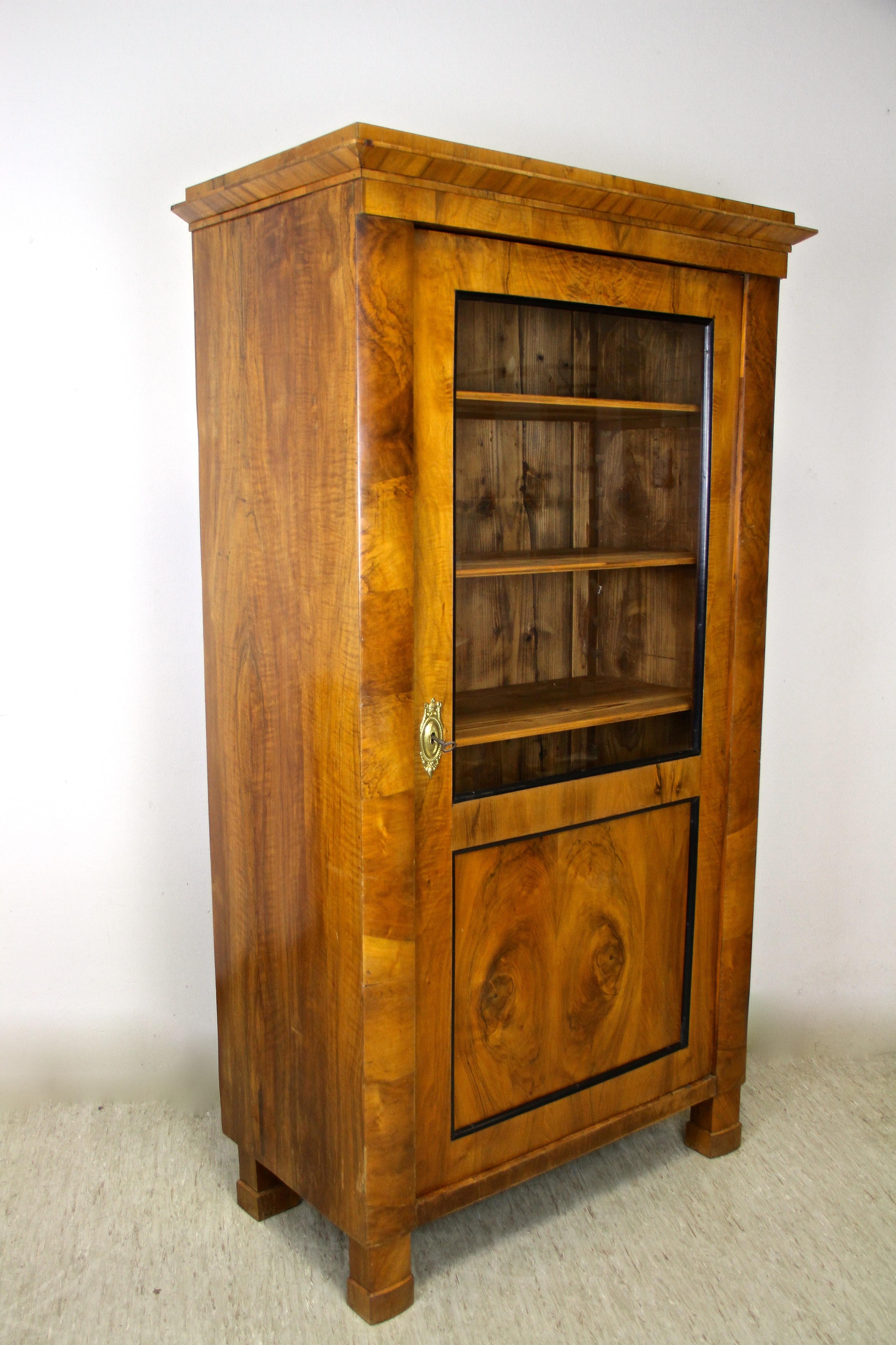 Lovely Biedermeier vitrine/ display cabinet from the renown period in Austria around 1840. A pleasant nutwood veneered 19th century cabinet with a perfect size - not too big and not too small. The door shows a big glass plate framed by ebonized bars
