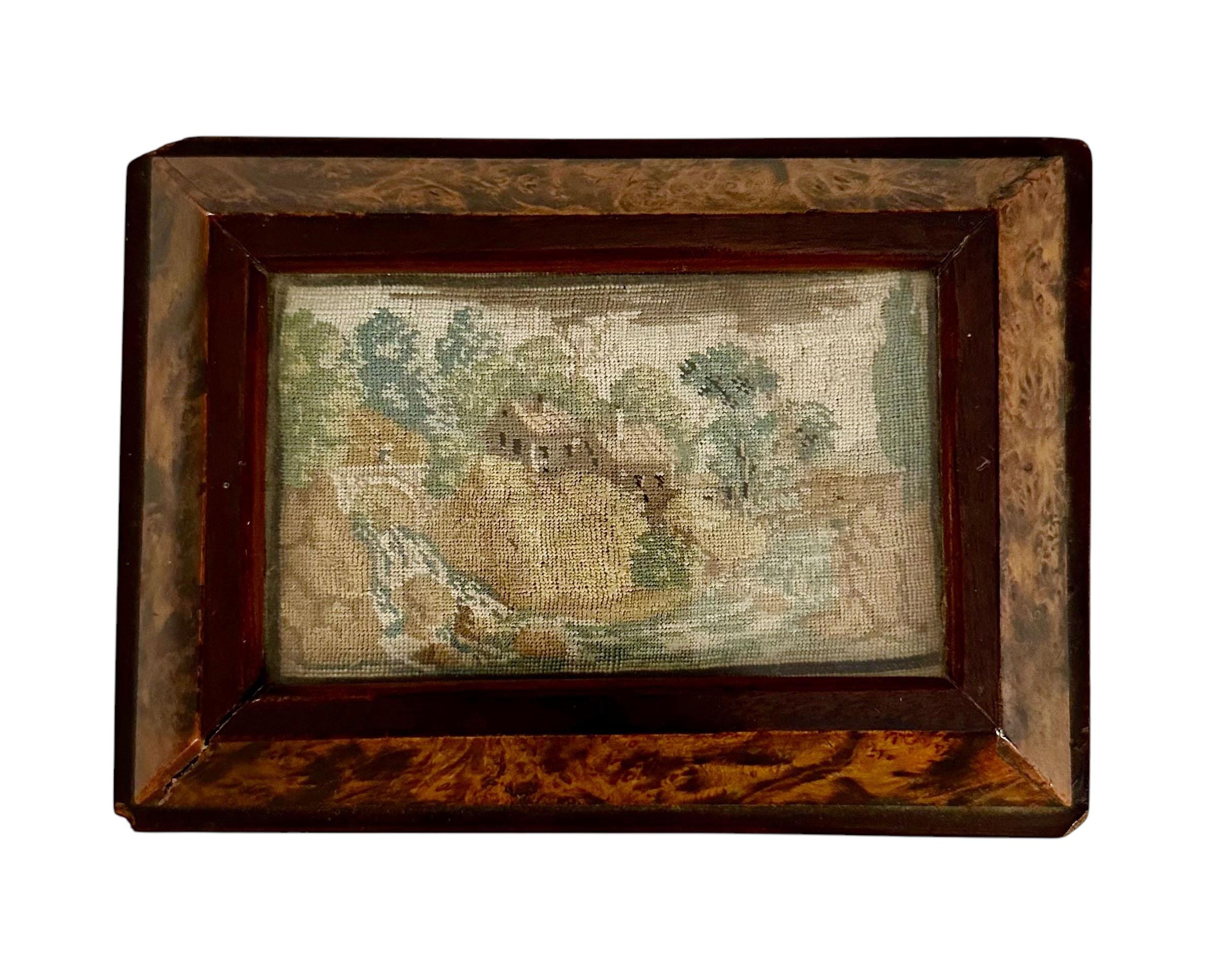 Caja Biedermeier alemana de los años 1830 a 1840 con inserción de petit point muy fino con cristal. La escena del petit point es de agua, árboles y dos casas. La caja tiene una base negra en la parte inferior. Tiene cerradura pero no llave.  