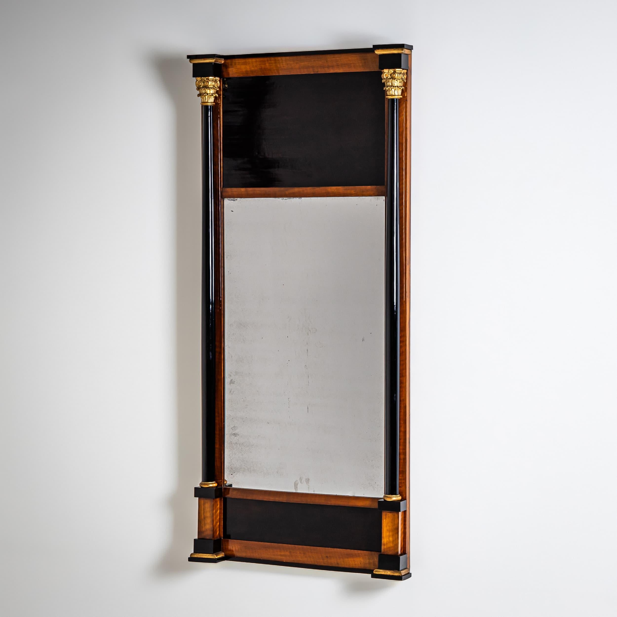 Elegante espejo de pared de formato esbelto y rectangular, fabricado hacia 1810. El marco está chapado en cálido cerezo y flanqueado por columnas completas ebonizadas con capiteles y bases patinados en oro. La placa de espejo original presenta los