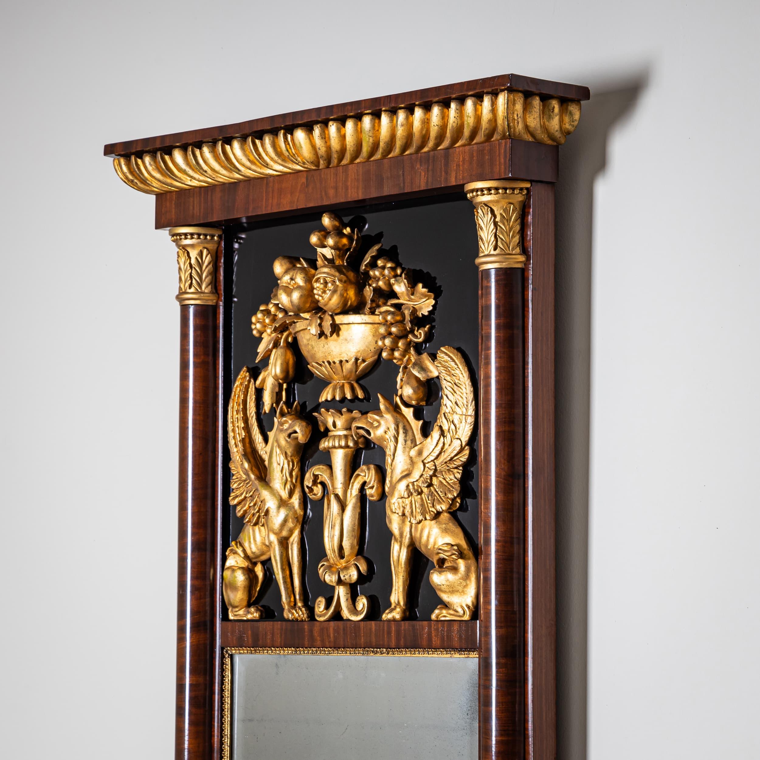 Biedermeier pier mirror veneered in mahogany with ebonized panel insets. Gilded stucco ornaments include rosettes, a fruit basket, and stylized griffins. The mirror is flanked by half-columns with gilt capitals and bases. It is crowned with a broad