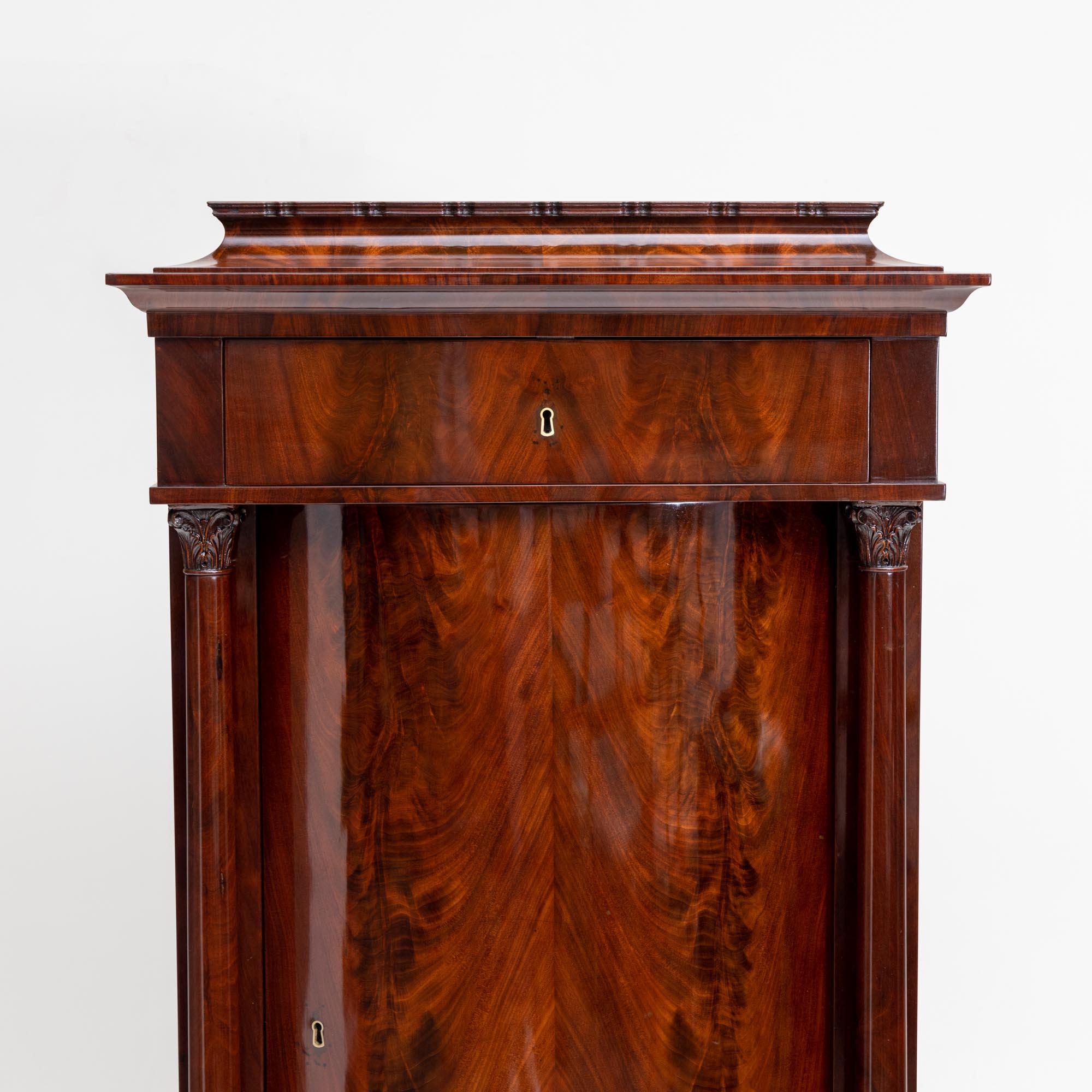 Mahogany veneered pillar cupboard with two drawers and a slightly convex door. The door is flanked by solid columns with leaf capitals. The cabinet stands on square feet and ends with a slightly raised central pedestal with a profiled edge. The