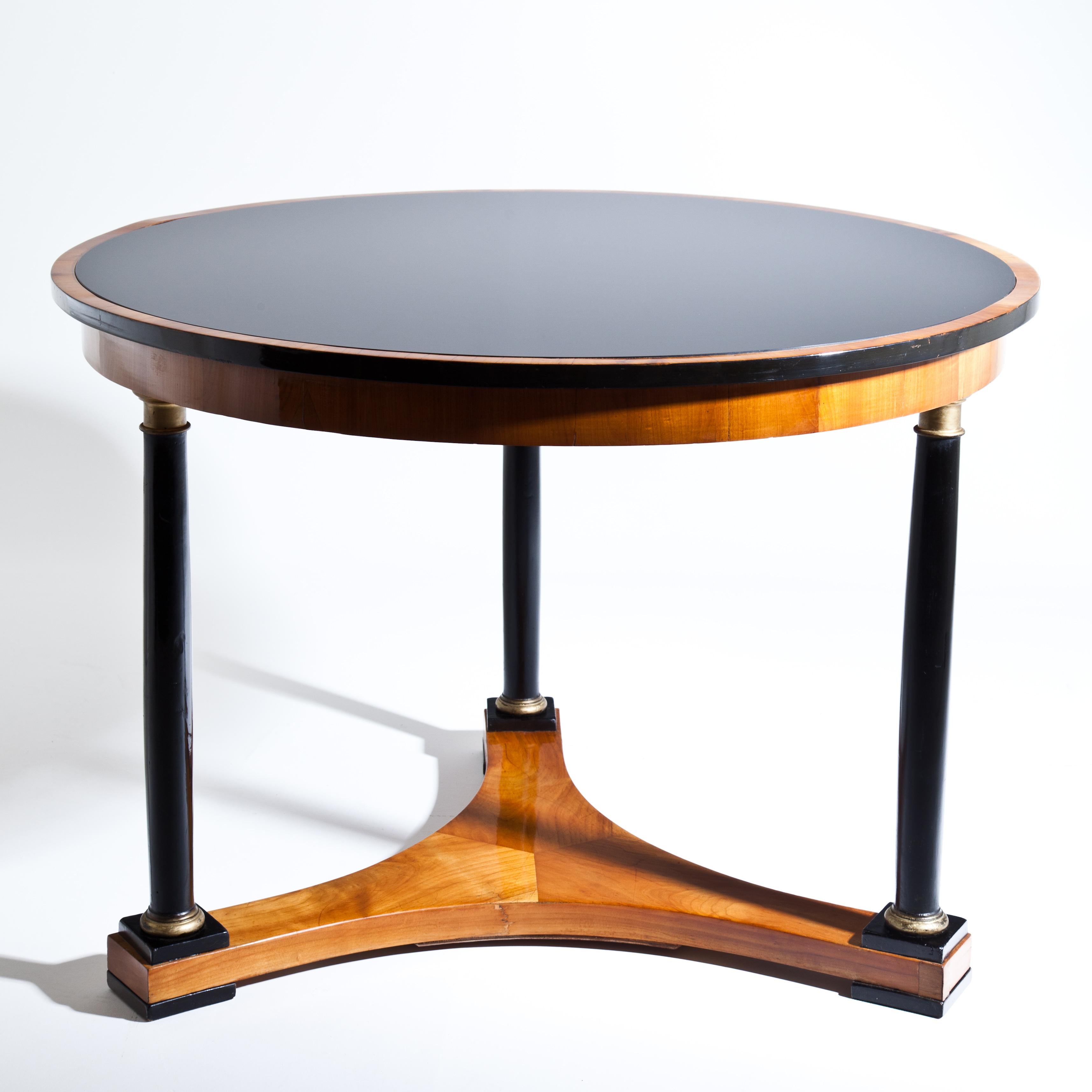 Biedermeier salon table in cherry, with round tabletop on three ebonized and partly gold-patinated column legs above a concave stand. The blackened tabletop with likewise ebonized edge has a recent glass top.
