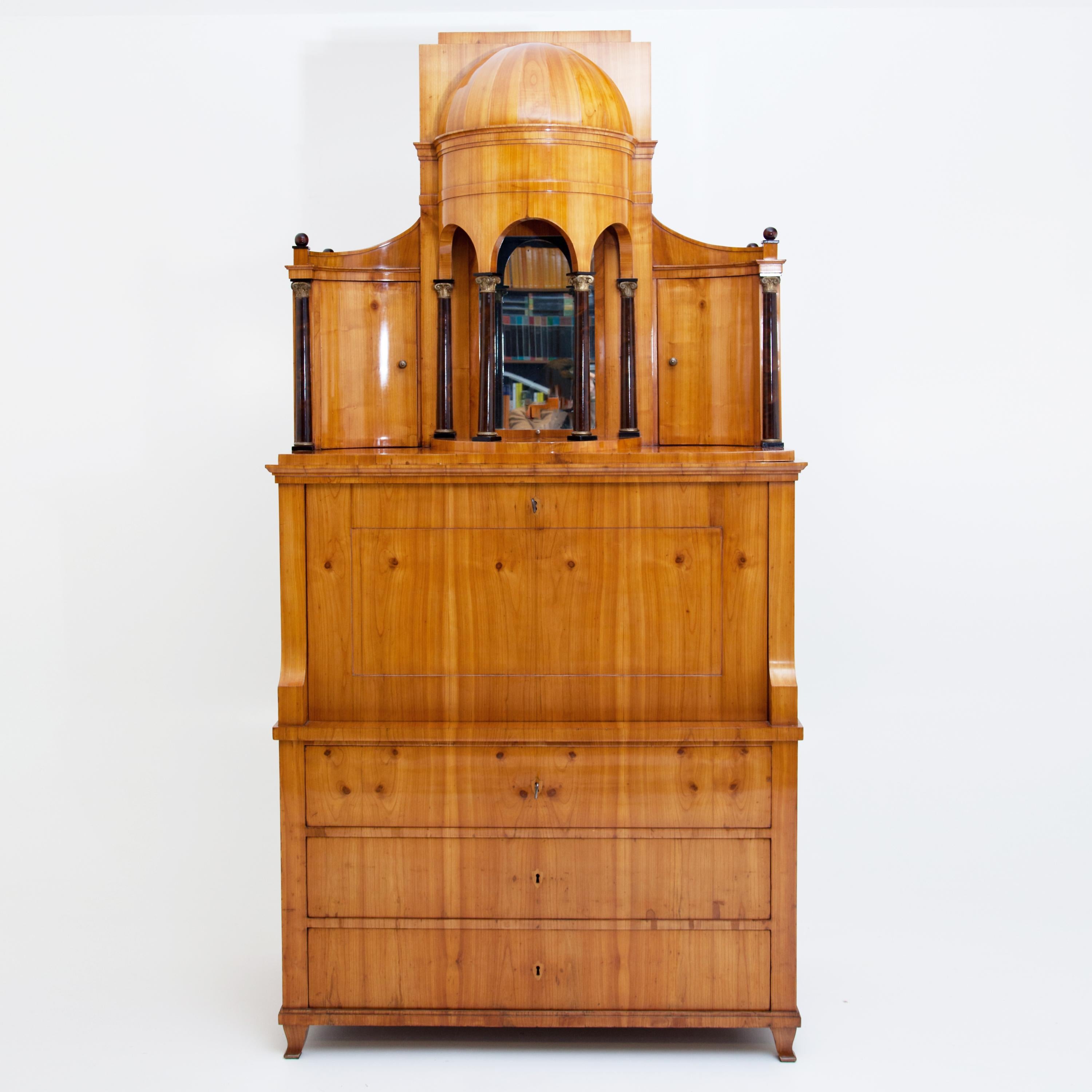 A secretaire with writing flap and an architectural top with partially gilded columns and half-dome rising above a three-drawered chest of drawers. Two concave side doors with slightly rising cornice lead to the high semi-dome with stepped top. The