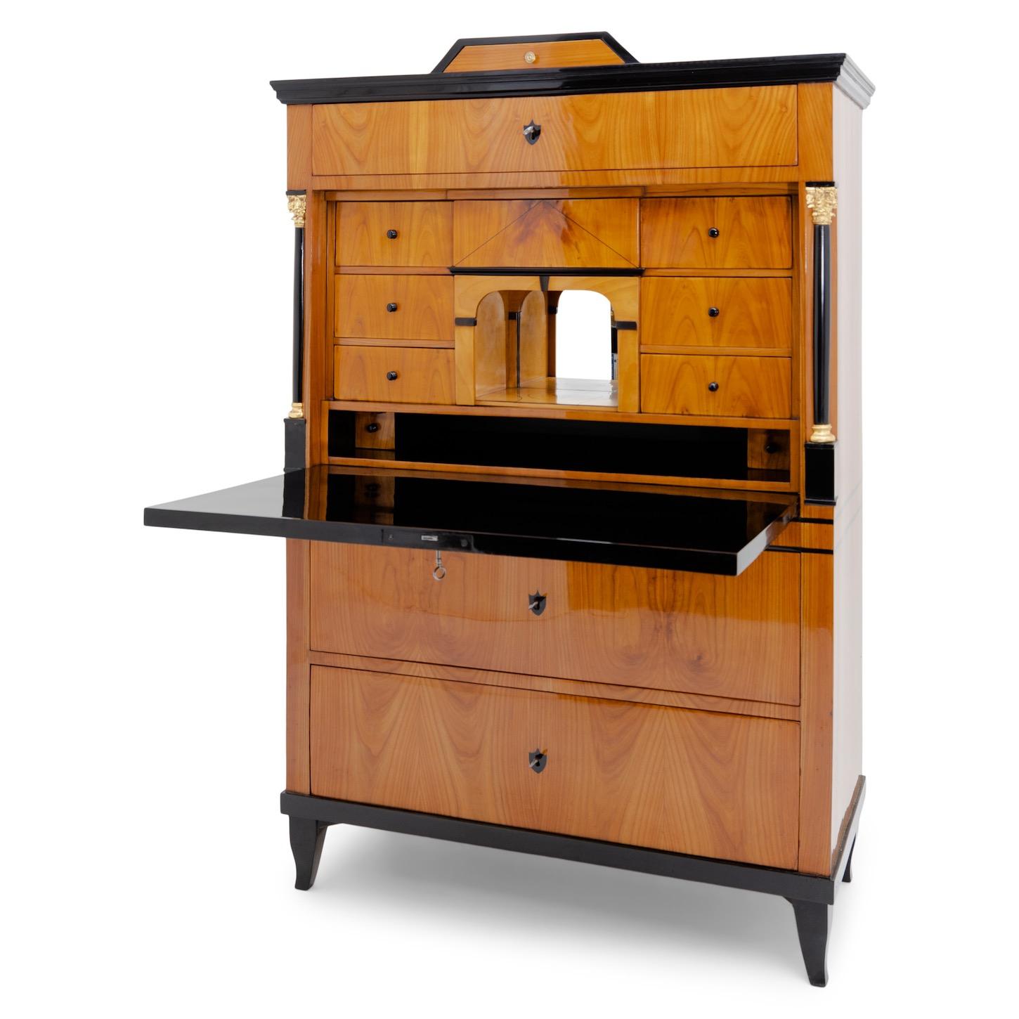 A cherry veneered secretaire standing on low ebonized S-legs, with a two-drawer chest of drawers and a rich interior. The secretaire is visually divided by a surrounding black double line. Ebonized and partly gilded columns with Corinthian capitals