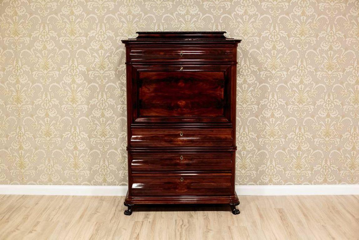 This intriguing piece of furniture is veneered with mahogany. It is dated circa 1880. The secretary has rounded shapes with rounded corners and cornices. The front is composed of four drawers and the lid. The fronts of the drawers are of a