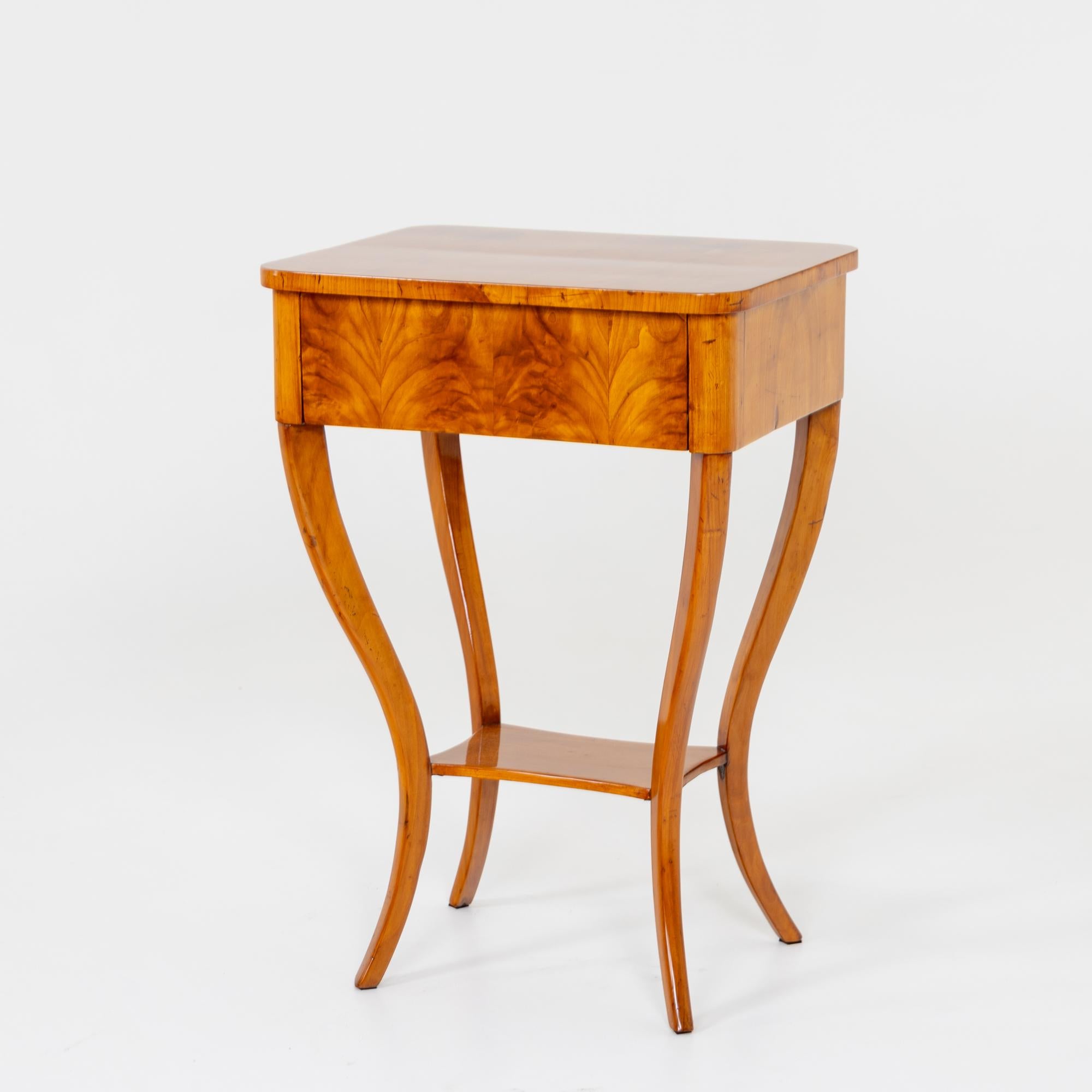 Small Biedermeier side table with high s-shaped legs and a small intermediate board. Above it rises the table top with a drawer and smooth frame. The table is veneered in cherry and has been polished by hand.